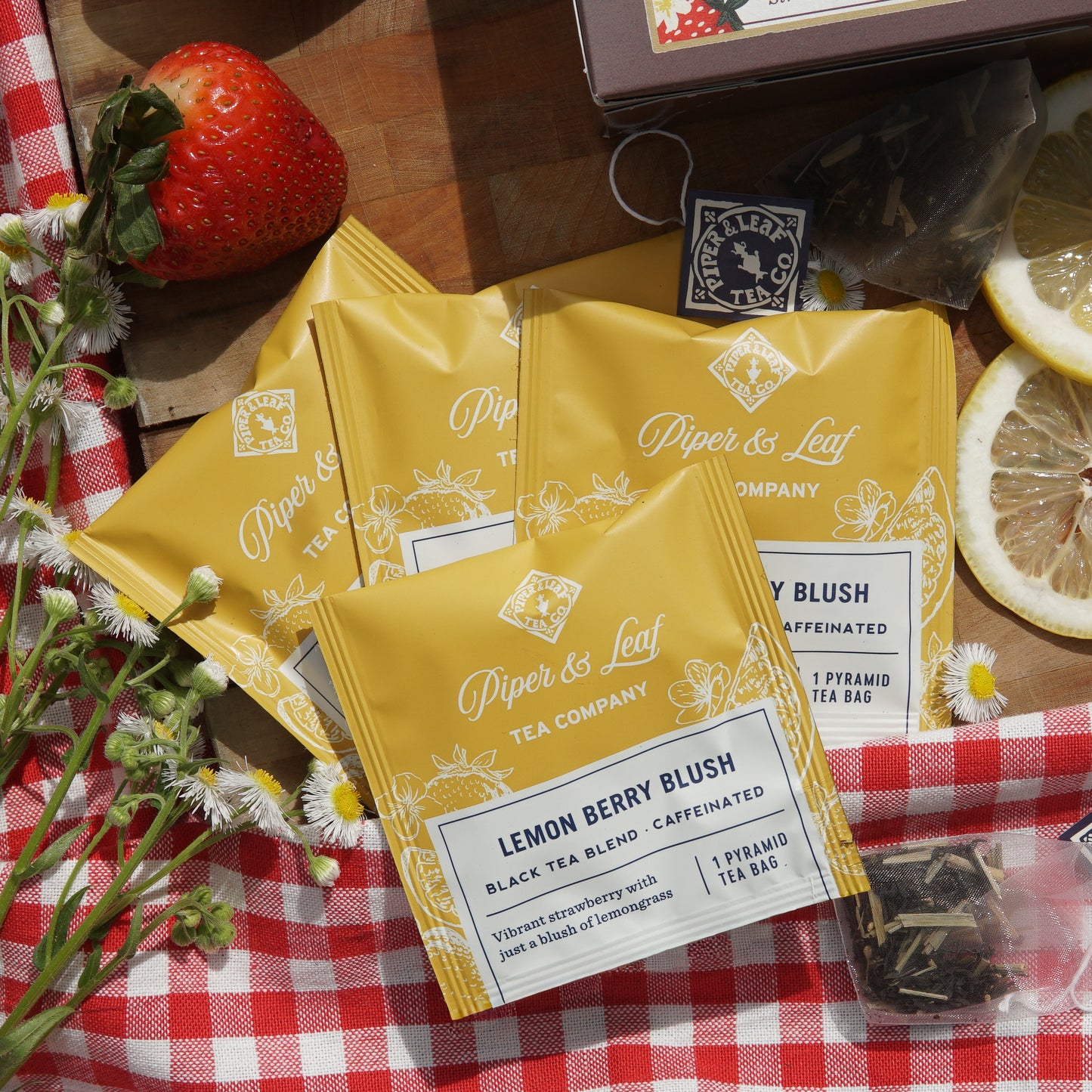 Lemon Berry Blush indiviually wrapped tea bags on top of a picnic blanket and wooden cutting board, surrounded by assorted lemons, strawberries, and wild flowers
