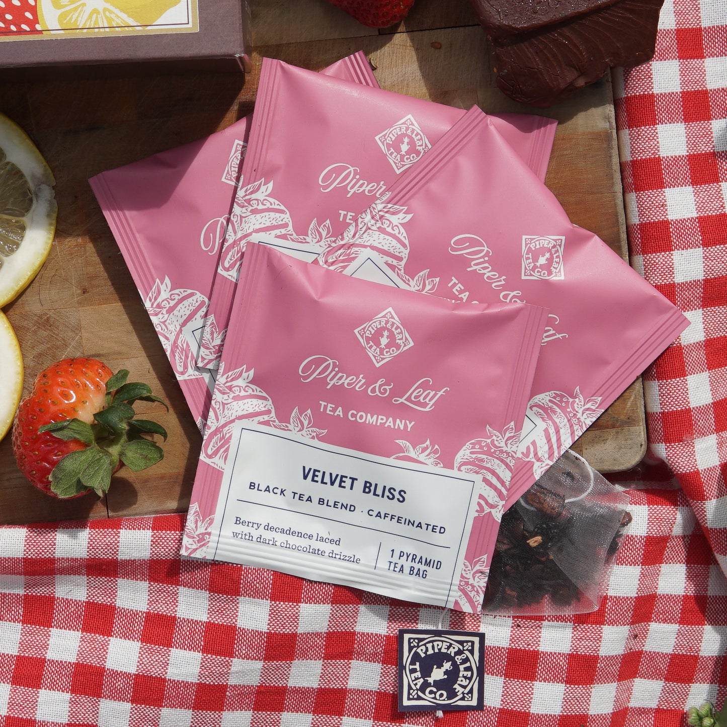 Velvet Bliss individually wrapped tea bags  on top of a picnic blanket and wooden cutting board  next to assorted strawberries and a chocolate chunk
