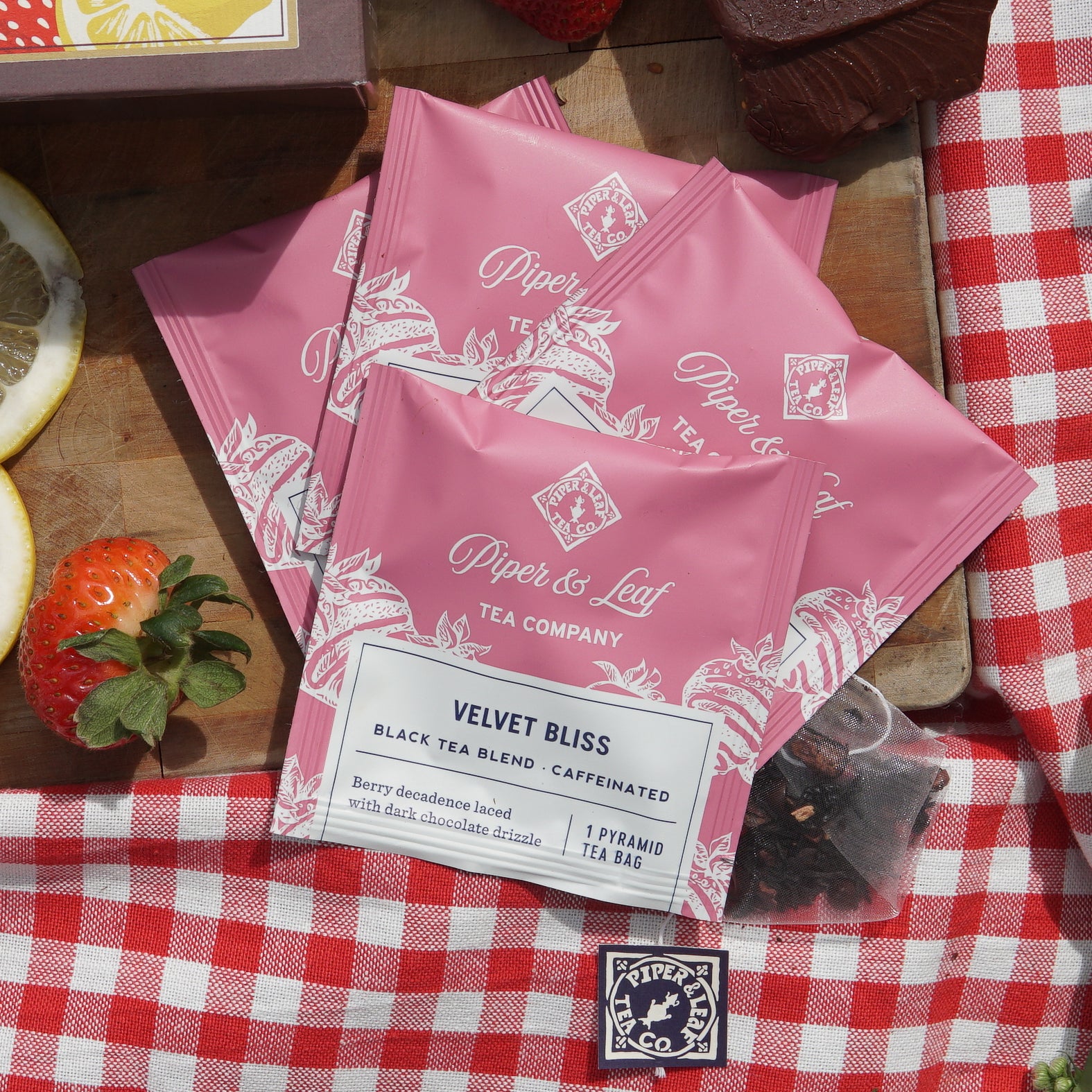 Velvet Bliss individually wrapped tea bags  on top of a picnic blanket and wooden cutting board  next to assorted strawberries and a chocolate chunk
