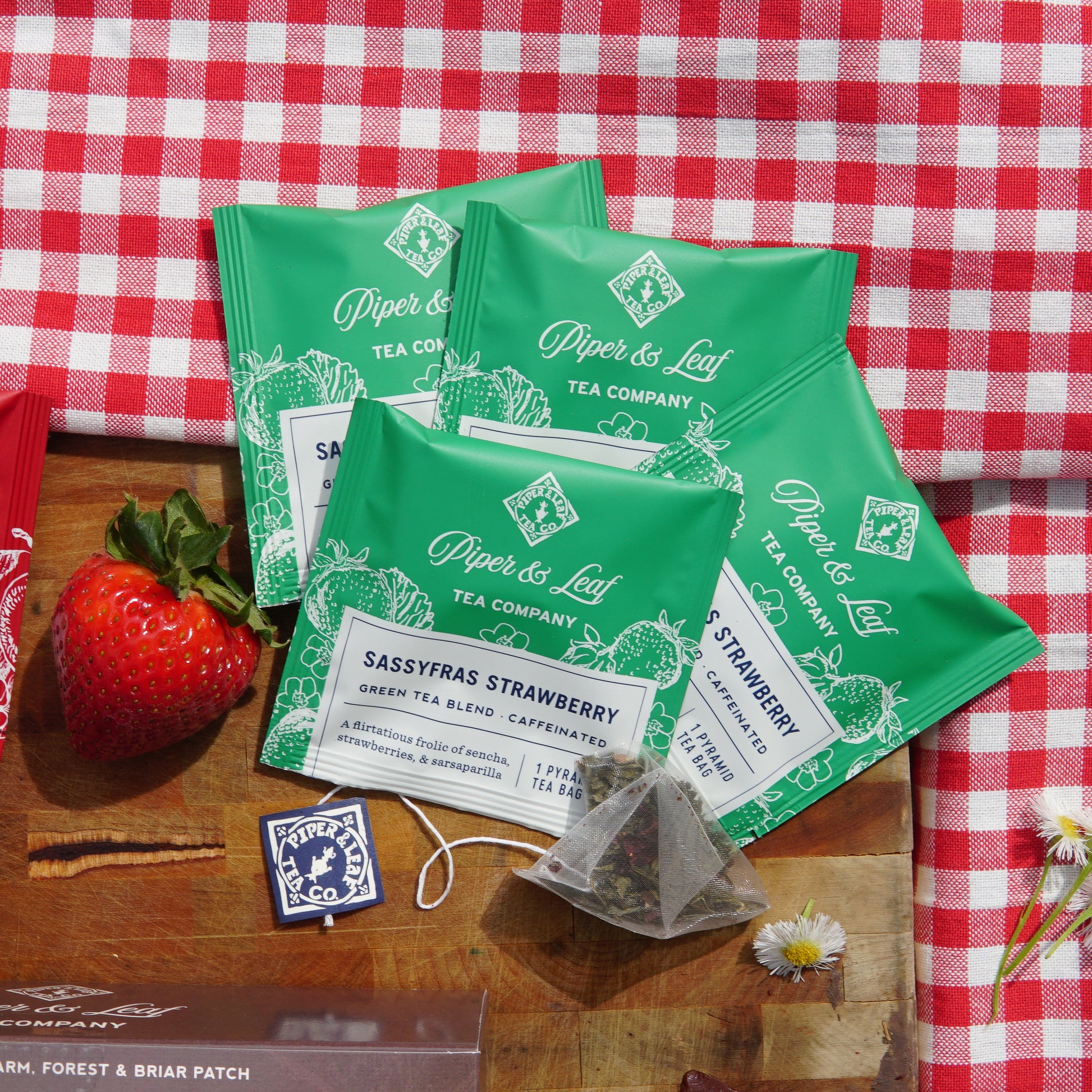 Sassyfras Strawberry individually wrapped tea bags on top of a picnic blanket and wooden cutting board next to a strawberry and wild flowers 