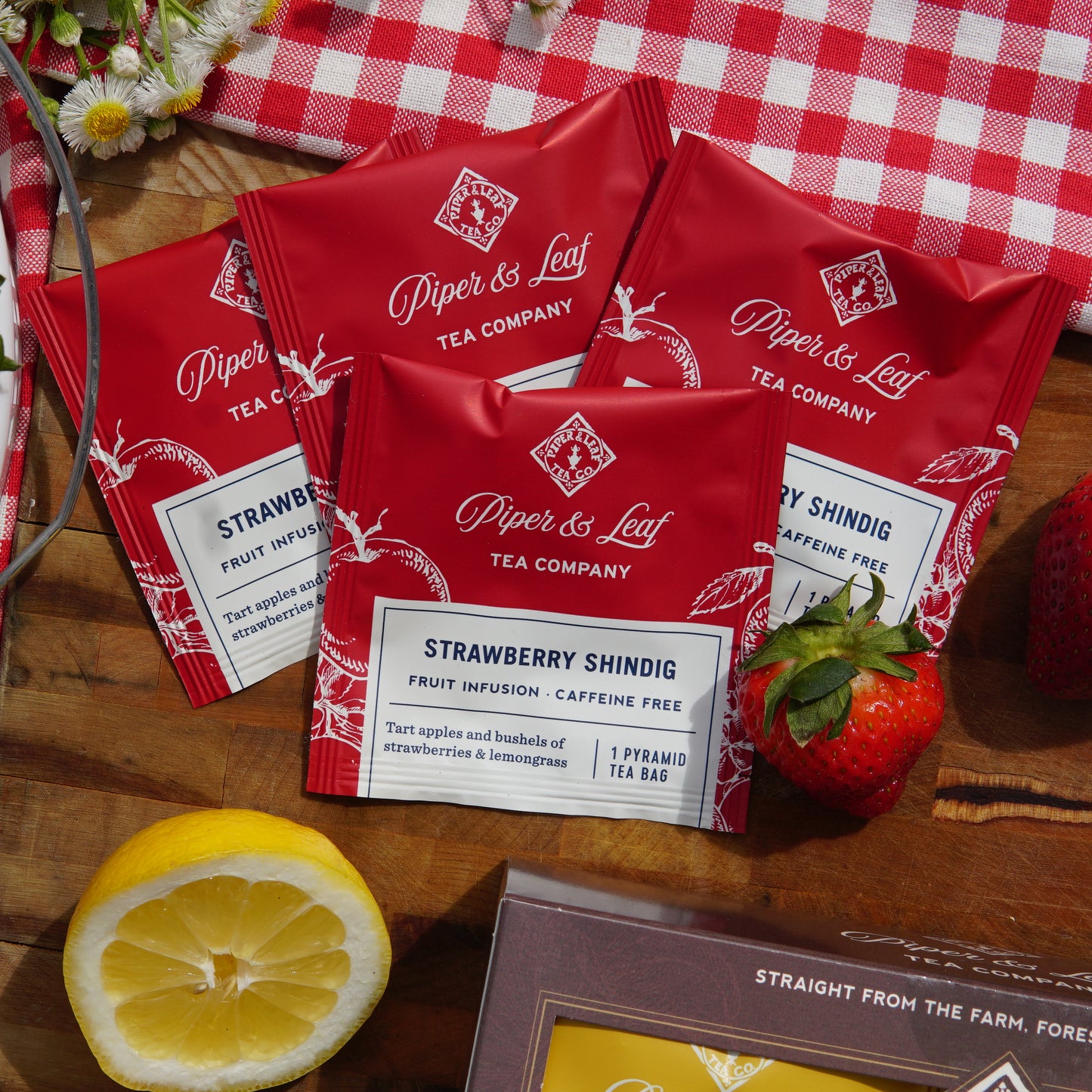 Strawberry Shindig individually wrapped tea bags on a picnic blanket and cutting board, surrounded by strawberries and a lemon slice 
