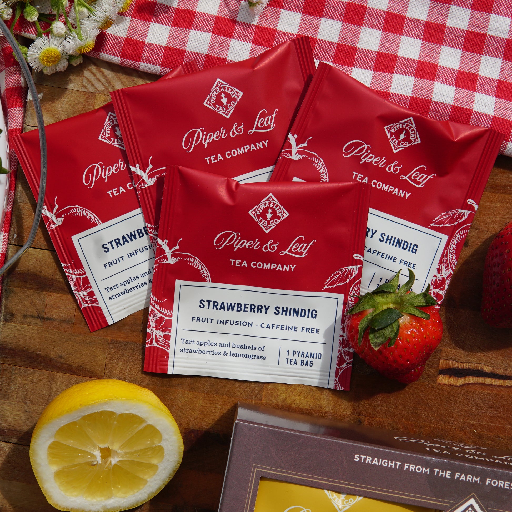 Strawberry Shindig individually wrapped tea bags on a picnic blanket and cutting board, surrounded by strawberries and a lemon slice 

