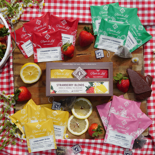 Strawberry Teas from Piper & Leaf Tea Co.  set on a picnic blanket with wooden cutting board. Featuring Strawberry Shindig, Sassyfras Strawberry, Lemon Berry Blush, and Velvet bliss. surrounded by assorted stawberries, lemons, and chocolate