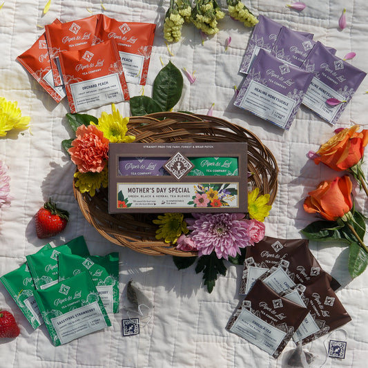 Piper & Leaf Tea Co.'s Mother's Day Special Tea Sampler features 16 individually wrapped tea bags in a gift box, beautifully arranged on a wicker tray with flowers, strawberries, and lemon atop a white patterned cloth.