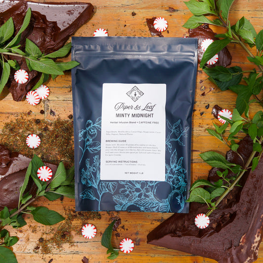 A Minty Midnight Pound Bag (190 Servings) by Piper & Leaf Tea Co. sits on a wooden surface, surrounded by fresh mint leaves, peppermint candies, and rich chocolate pieces.