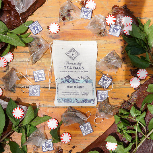 A pouch of Piper & Leaf Tea Co. "Minty Midnight 9ct Tea Bags in Muslin" is placed on a wooden surface, surrounded by creamy cocoa pieces, mint leaves, chocolate, and peppermint candies.