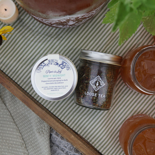 A tray holds yellow flowers, a mint plant, several mason jars of iced tea—including one with Piper & Leaf Tea Co.’s Minty Midnight Glass Jar of Loose Leaf (30 servings)—a candle, and a tin atop a striped cloth.