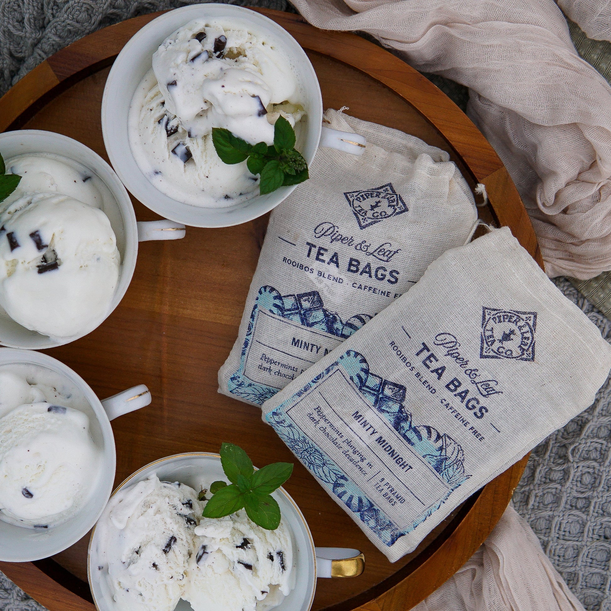 Four cups of mint chocolate ice cream with fresh mint are on a wooden tray next to two Piper & Leaf Tea Co. Minty Midnight 9ct Tea Bags in Muslin; gold spoons and a plate are placed nearby.