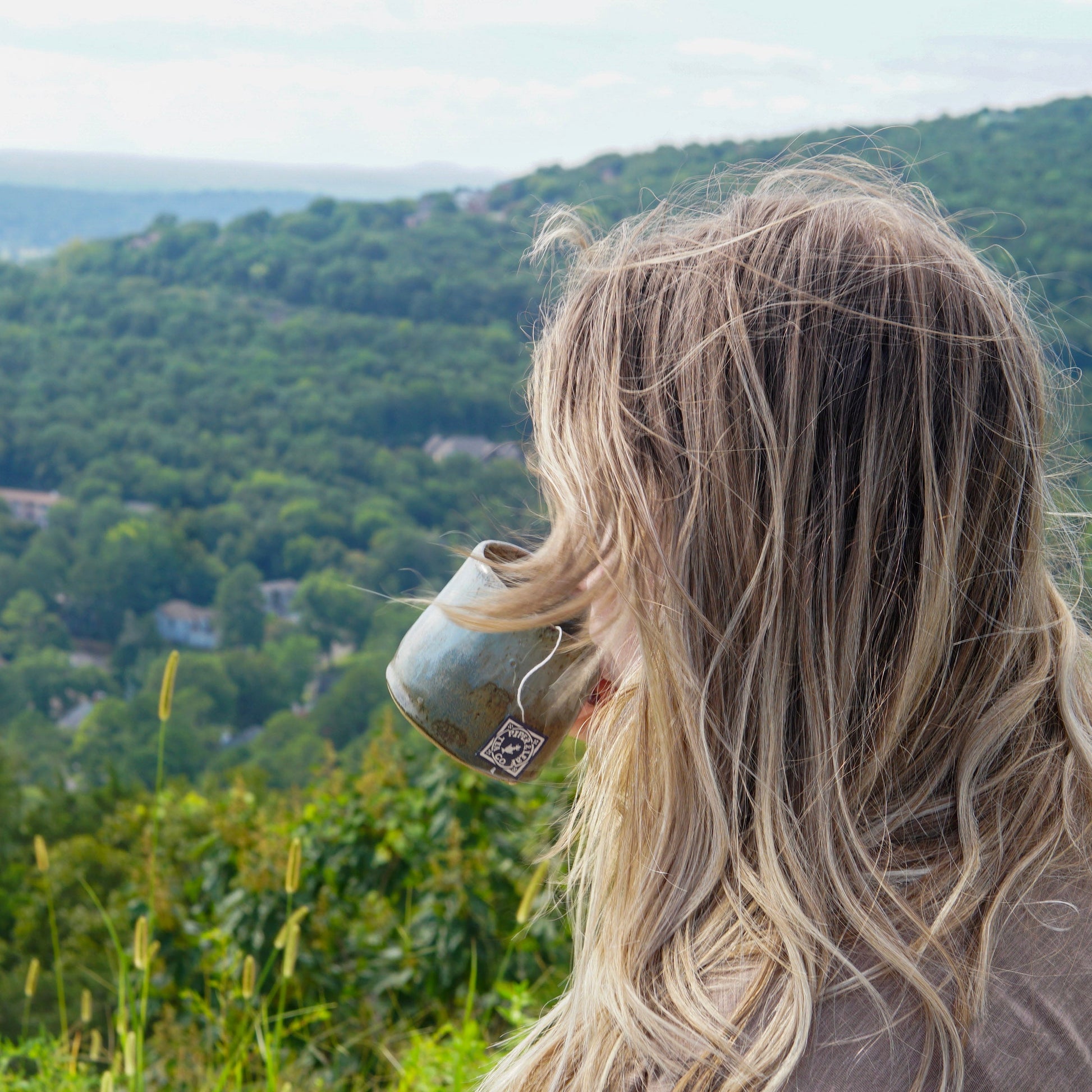 A person with long blonde hair drinks Piper & Leaf Tea Co.'s Mental Clarity from a mug, enjoying mood balance and stress resilience while overlooking a green, tree-covered landscape and distant hills.