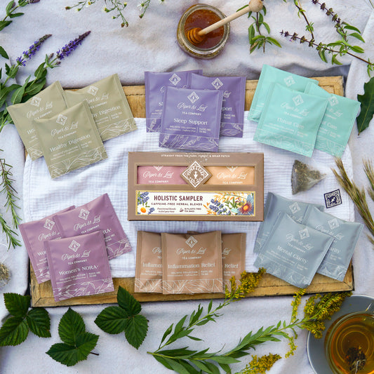 A Piper and Leaf Tea Co. Holistic Collection Sampler box of 18 tea bags surrounded by individual tea packets, a jar of honey, and fresh herbs on a wooden tray.
