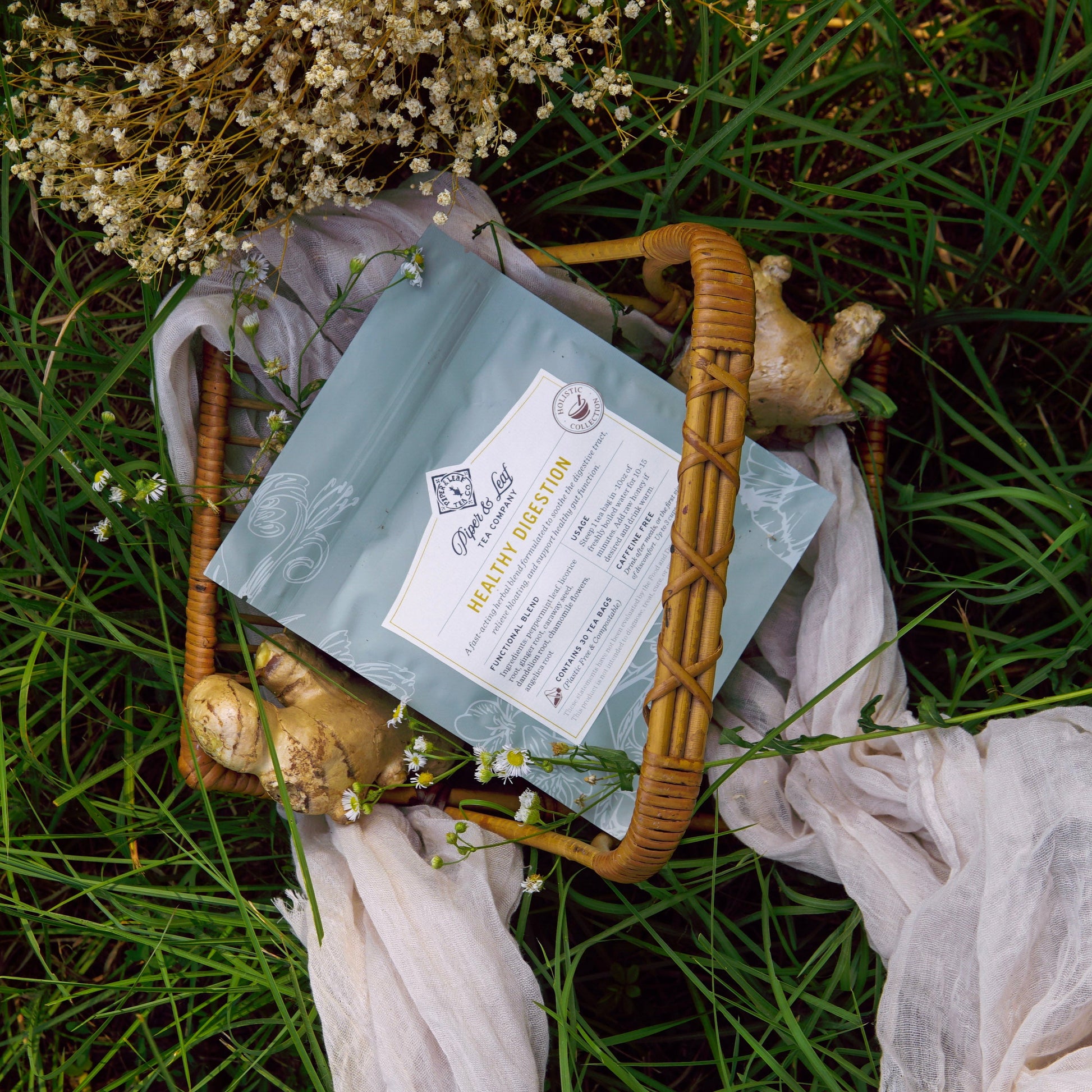 A wicker basket with a pouch of Piper & Leaf Tea Co.’s Healthy Digestion, surrounded by ginger, white flowers, and a white cloth on grass—a soothing herbal blend for digestive support and relief from bloating.