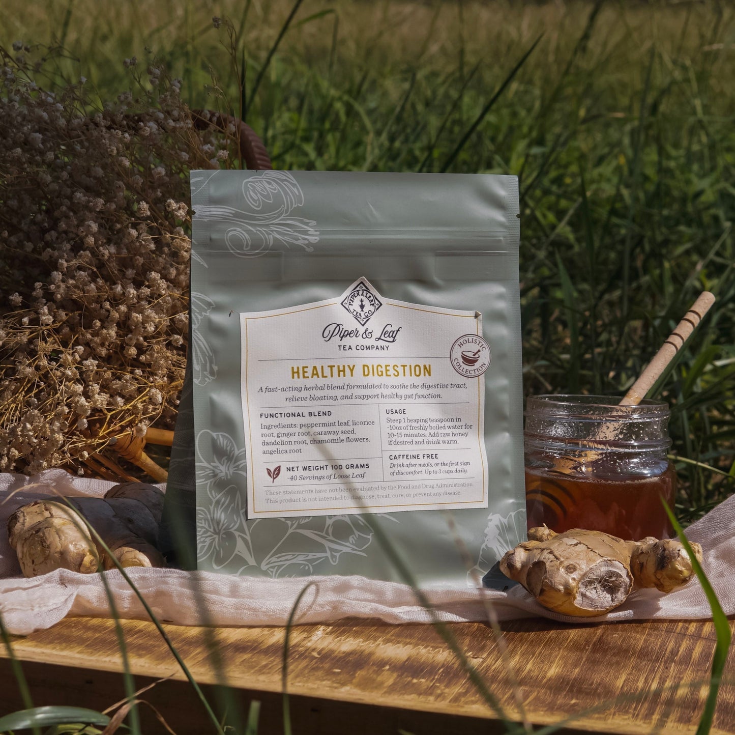 A bag of Piper & Leaf Tea Co. Healthy Digestion sits on a wooden tray outdoors, surrounded by ginger roots, honey, a honey dipper, and dried flowers—an herbal blend designed for gentle digestive support.