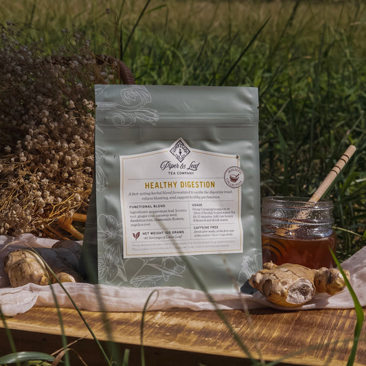 A bag of Piper & Leaf Tea Co. Healthy Digestion sits on a wooden tray outdoors, surrounded by ginger roots, honey, a honey dipper, and dried flowers—an herbal blend designed for gentle digestive support.