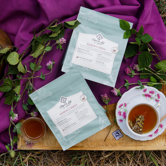 Two packages of Piper & Leaf Tea Co.’s Women’s NORA+ sit on a picnic blanket amid flowers and leaves, with a cup and glass of brewed tea—an inviting setup that highlights women’s wellness and hormone balance support.