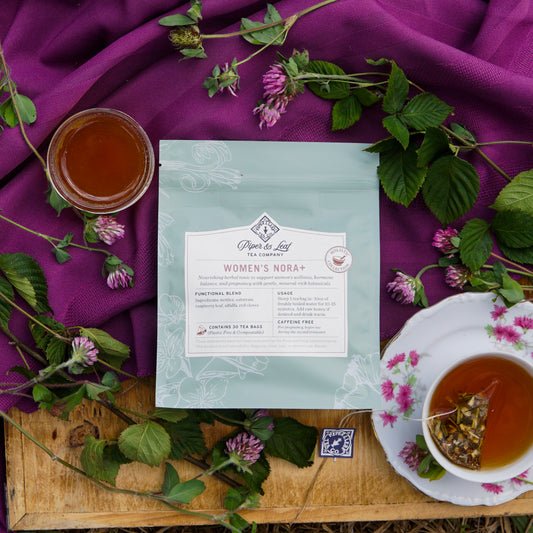A package of Piper & Leaf Tea Co.’s Women’s NORA+ tea is displayed with a tea cup, a glass cup, and green leaves on a purple cloth atop a wooden surface.