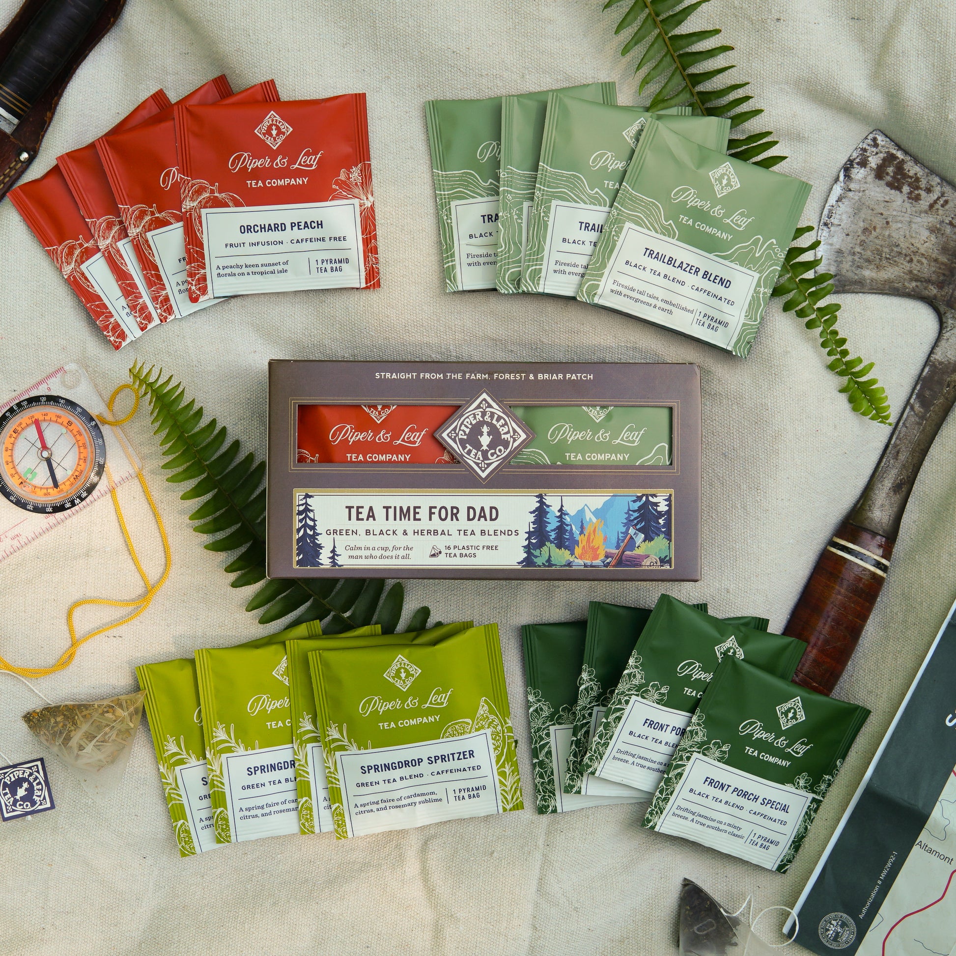 A flat lay featuring Piper & Leaf Tea Co.'s "Tea Time For Dad" box with 16 individually wrapped tea bags, a compass, axe, scissors, and garden trowel on fabric—a unique Father's Day gift for tea-loving dads.