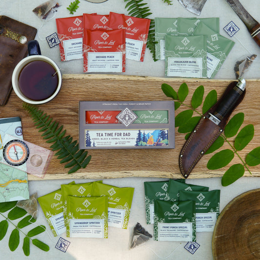 A box of 16 individually wrapped "Tea Time For Dad" tea bags by Piper & Leaf Tea Co. sits on a wooden board with a cup of tea, map, compass, leaves, and knife—an ideal Father’s Day tea gift.