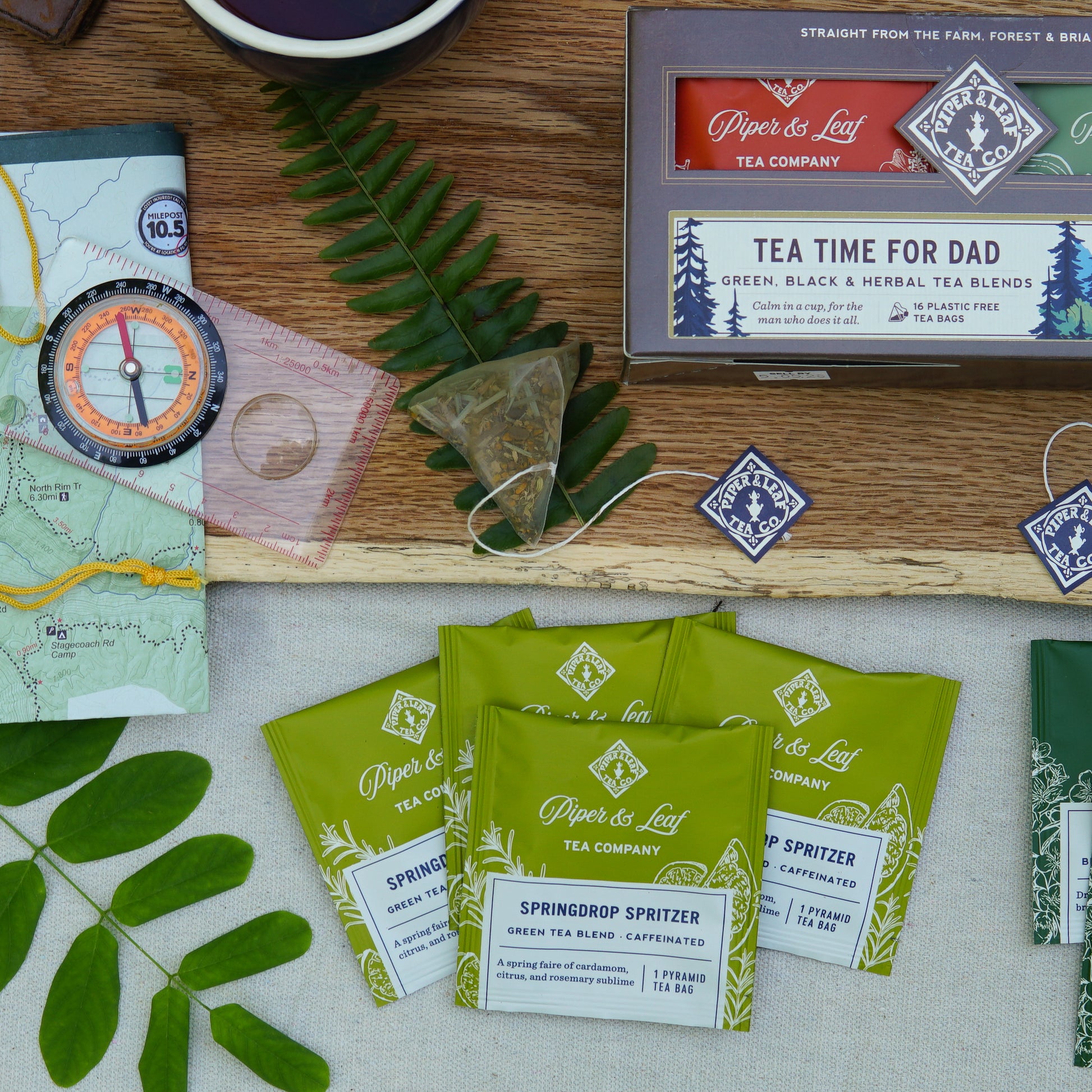 A flat lay featuring Piper & Leaf Tea Co.'s "Tea Time For Dad" box of 16 individually wrapped tea bags, a tea infuser, mug, compass, map, and greenery—ideal inspiration for the perfect Father's Day gift.