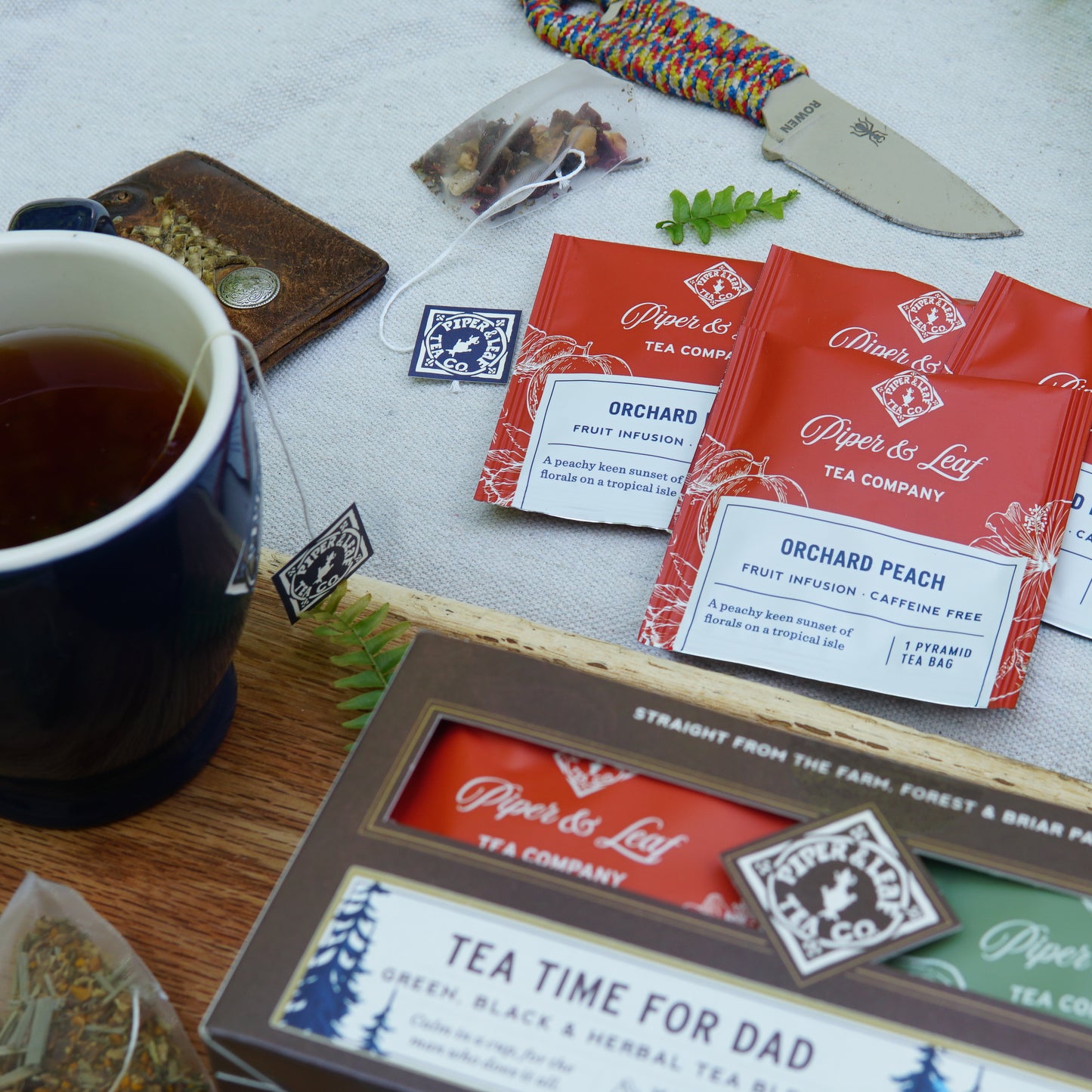 A cup of tea, Piper & Leaf Tea Co.'s "Tea Time For Dad" box of 16 individually wrapped tea bags, a knife, loose tea leaves, and a braided bracelet are artfully arranged on a white surface.