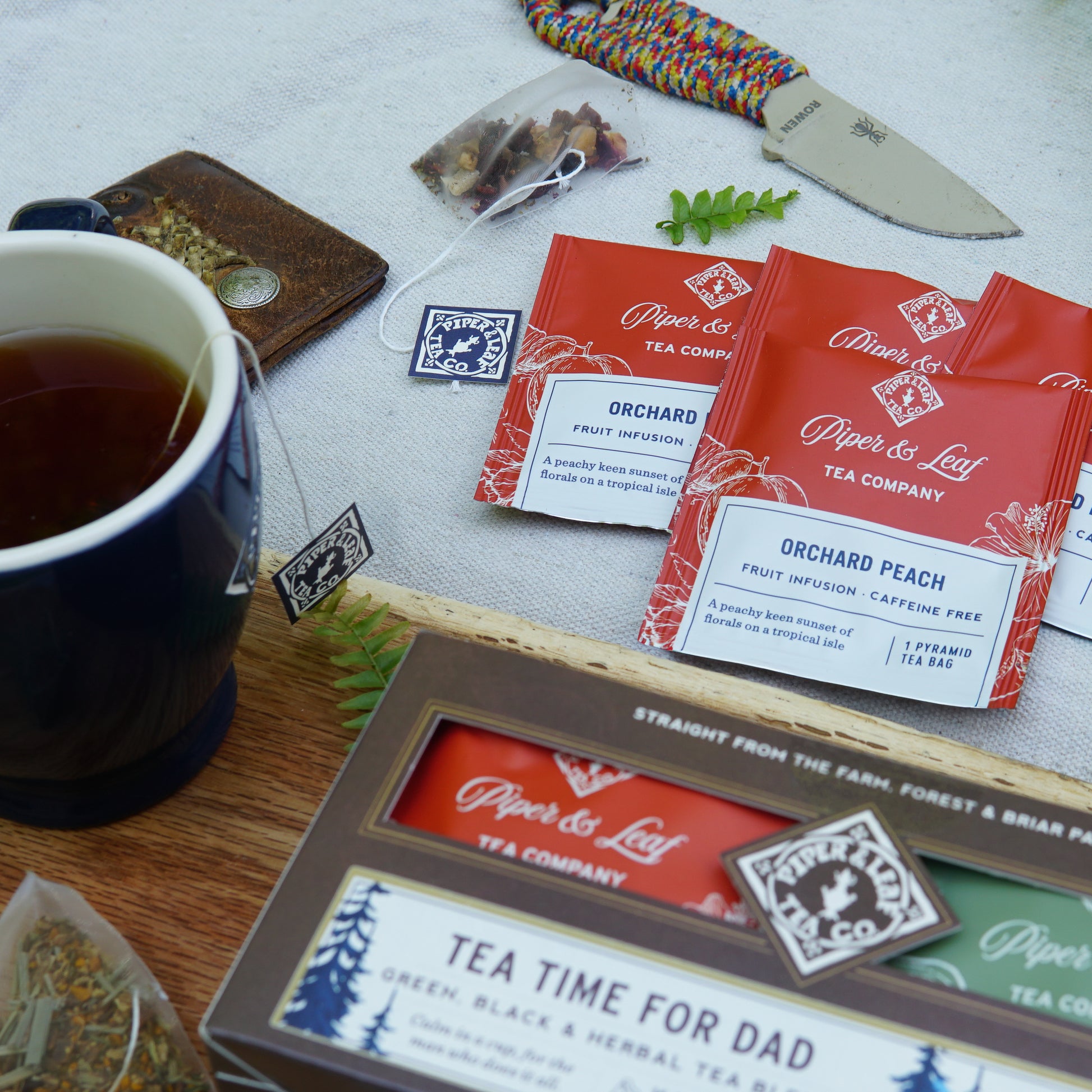 A cup of tea, Piper & Leaf Tea Co.'s "Tea Time For Dad" box of 16 individually wrapped tea bags, a knife, loose tea leaves, and a braided bracelet are artfully arranged on a white surface.