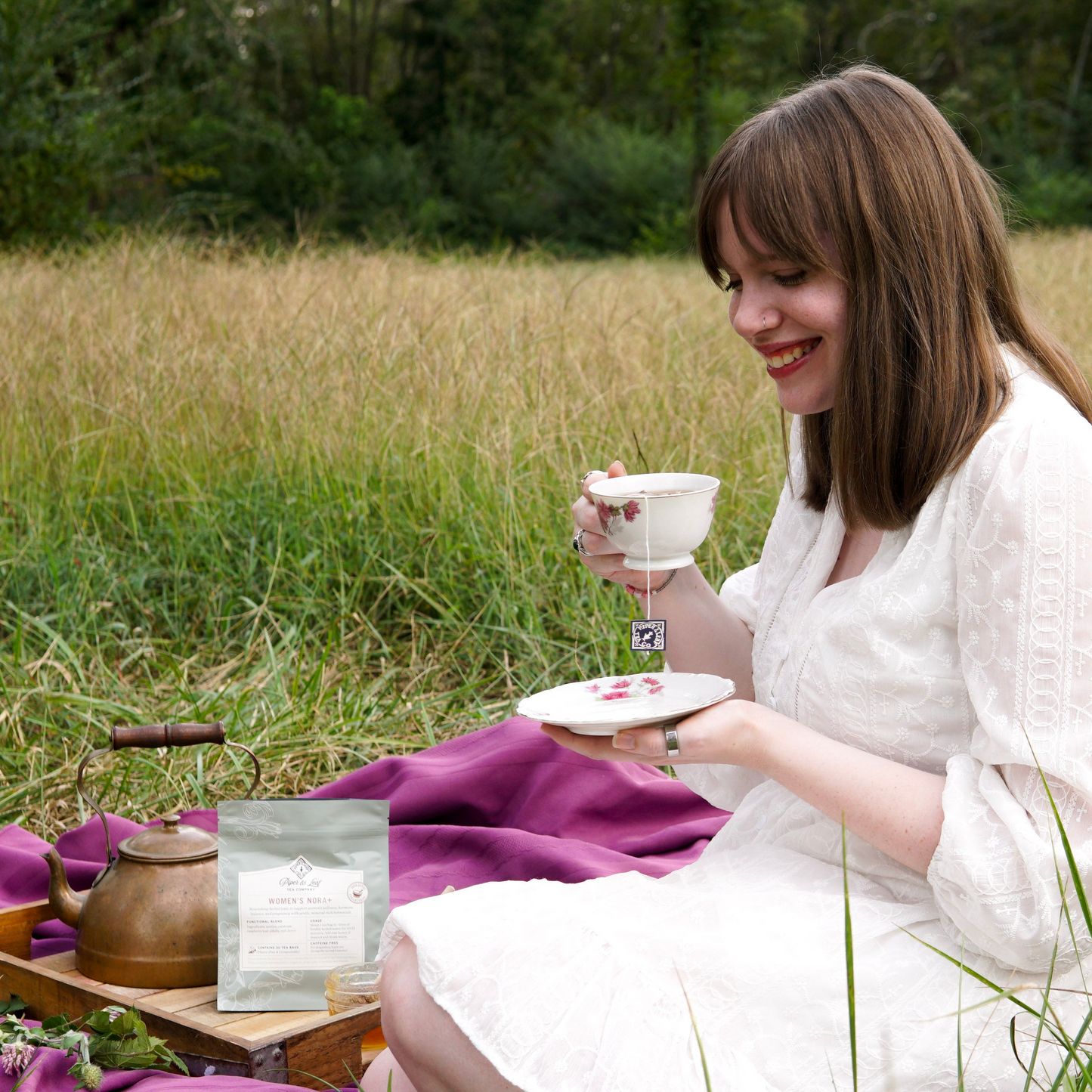 A woman in a white dress sits on a blanket in a grassy field, smiling with a teacup and saucer. In front of her are a teapot and Piper & Leaf Tea Co.'s Women’s NORA+ tea, which supports women's wellness and hormonal balance.