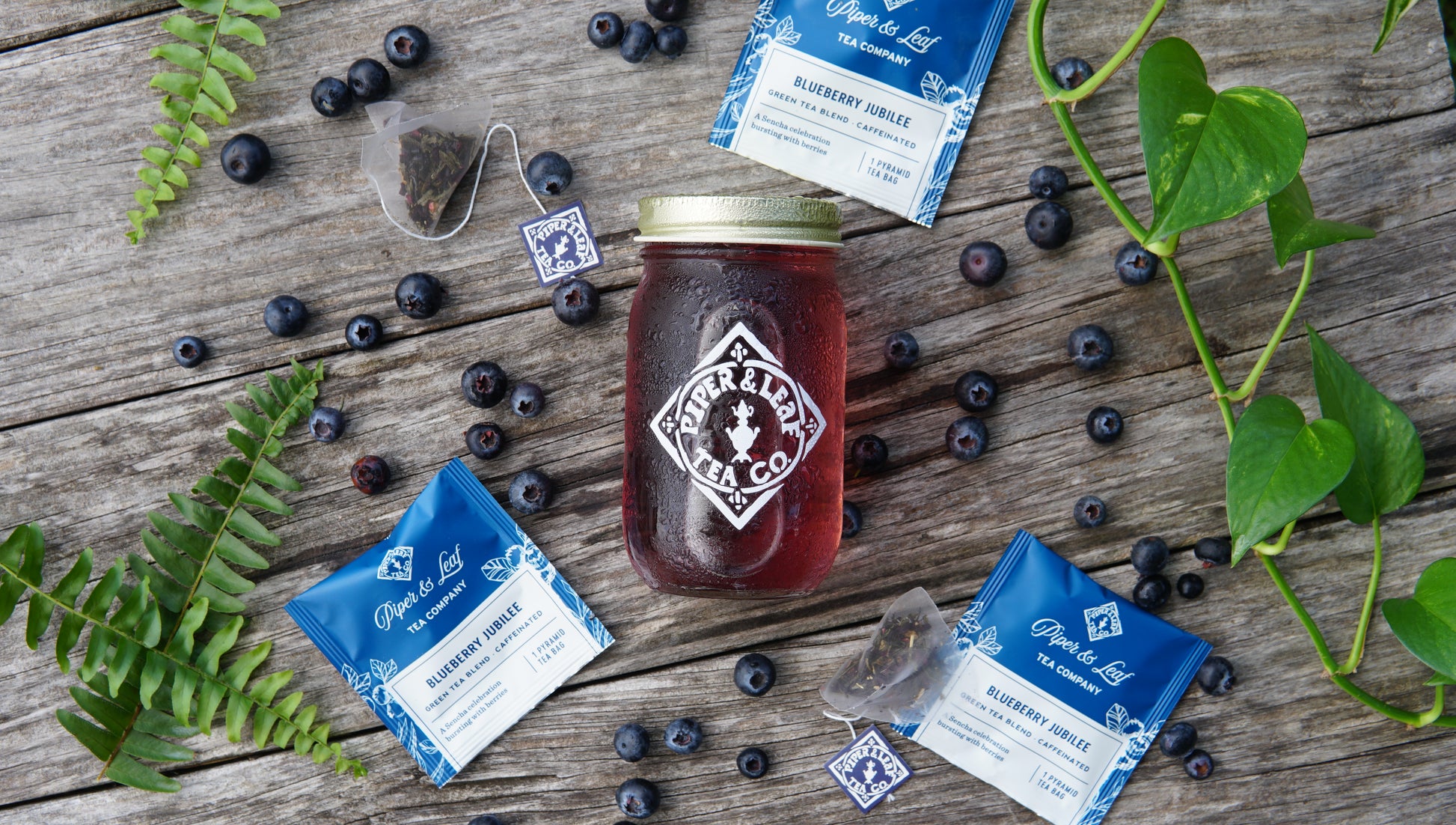 A jar of red iced tea from Piper & Leaf Tea Co.'s Blueberry Jubilee—Box of 12 Sachets—rests on wood, surrounded by tea bags, blueberry packets, fresh blueberries, green leaves, and a touch of Sencha for a refreshing limited edition blend.