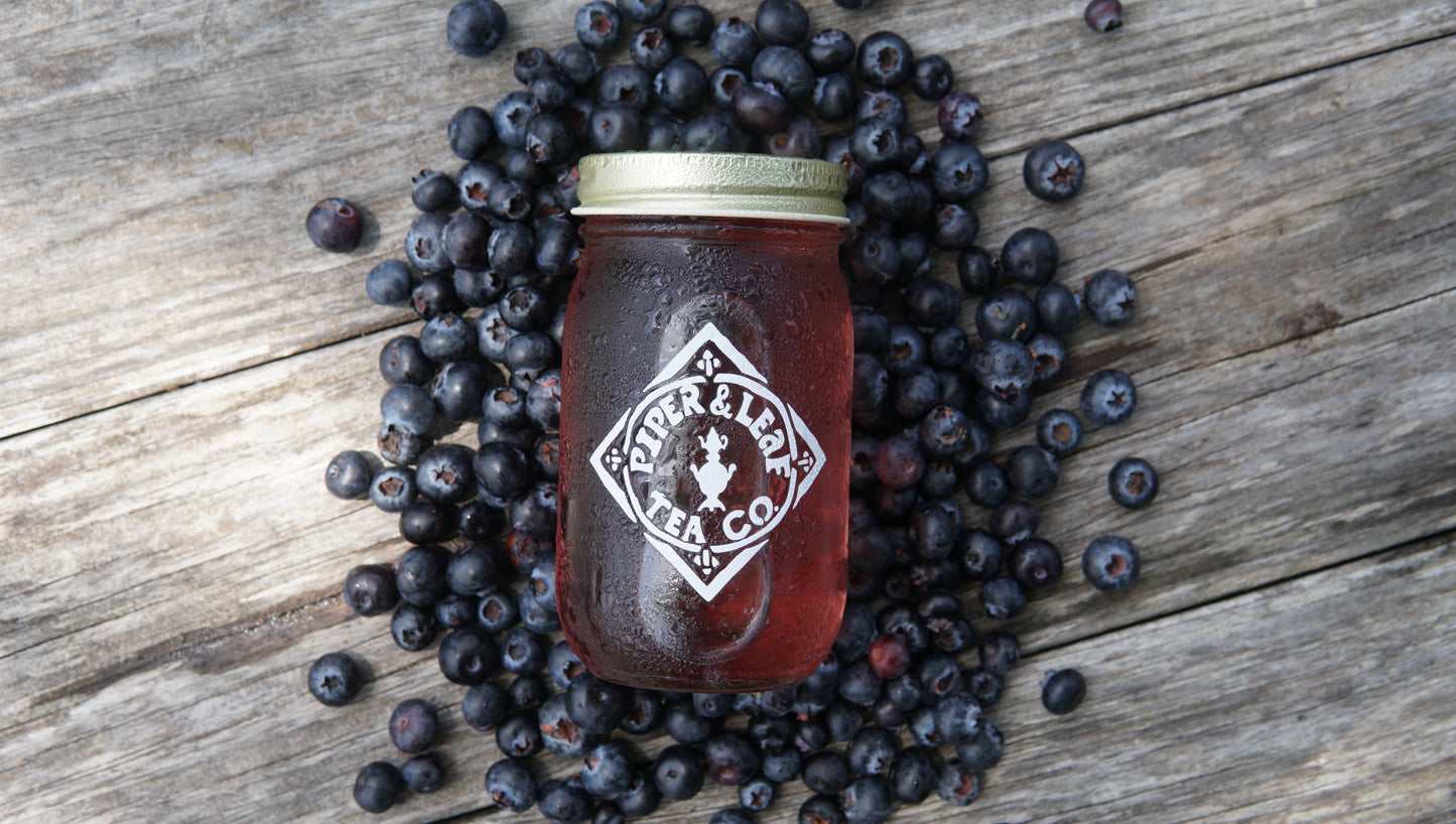 A mason jar filled with red liquid, labeled “Piper and Leaf Tea Co.,” sits on a wooden surface surrounded by scattered blueberries, showcasing their Blueberry Jubilee 40 Serving Loose Leaf tea.