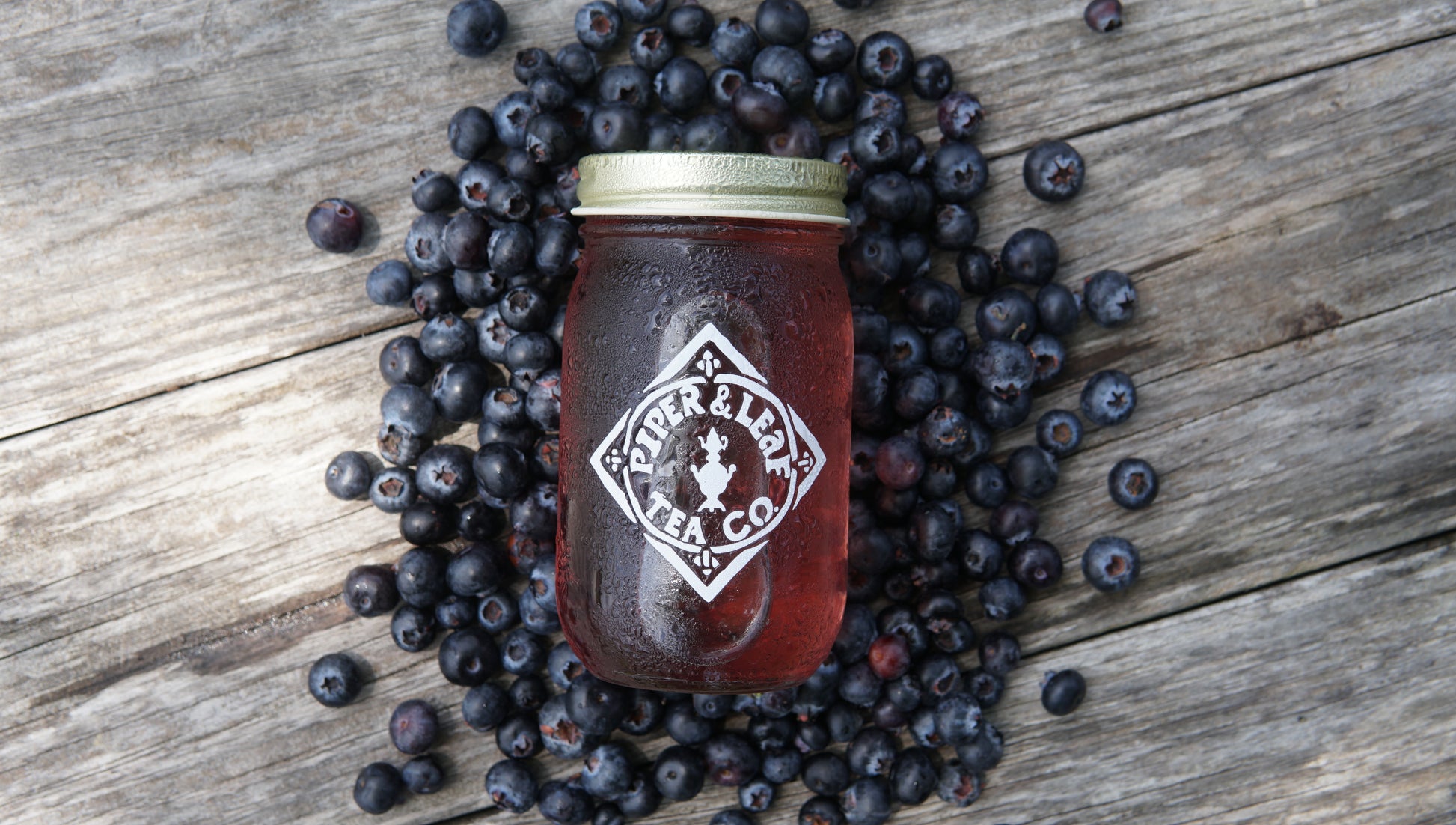 A mason jar filled with red liquid, labeled “Piper and Leaf Tea Co.,” sits on a wooden surface surrounded by scattered blueberries, showcasing their Blueberry Jubilee 40 Serving Loose Leaf tea.