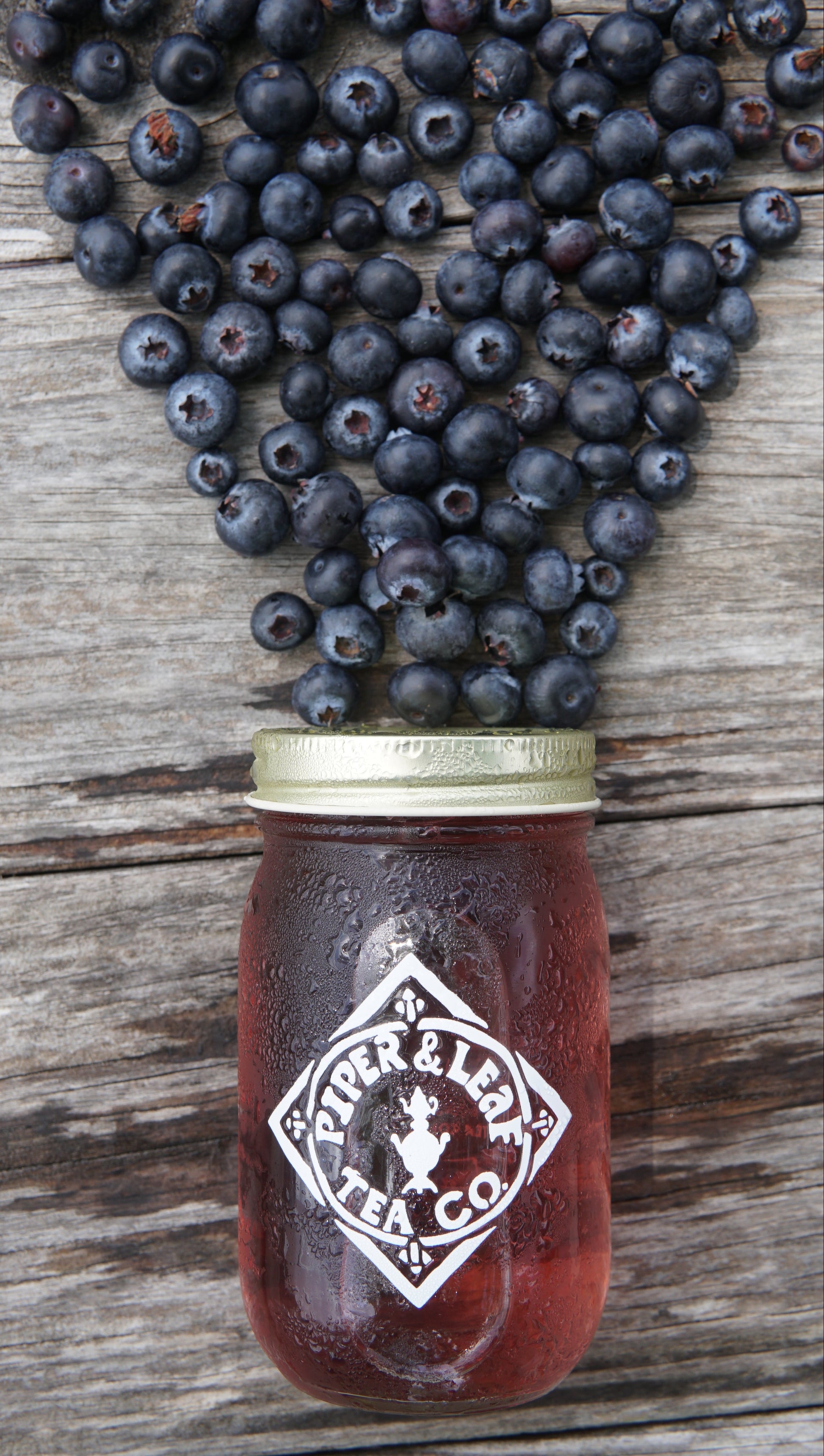 A mason jar of tea labeled "Piper and Leaf Tea Co." sits on wood, with blueberries above as if pouring in, highlighting their Blueberry Jubilee 40 Serving Loose Leaf limited edition blend.