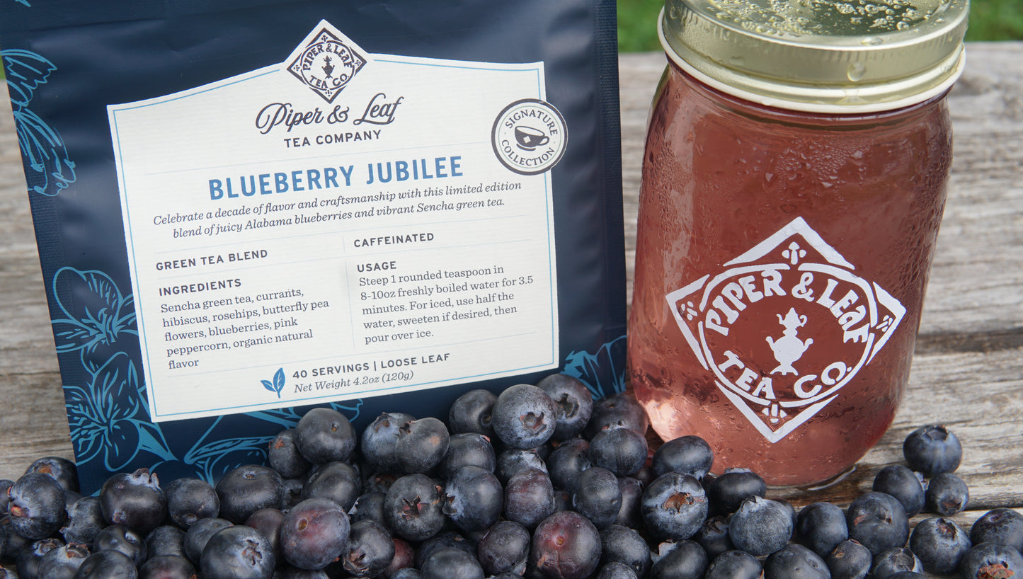 A package of Piper and Leaf Tea Co. Blueberry Jubilee 40 Serving Loose Leaf, a glass jar with their logo filled with the limited edition blend, and freshly picked blueberries arranged on a wooden surface.