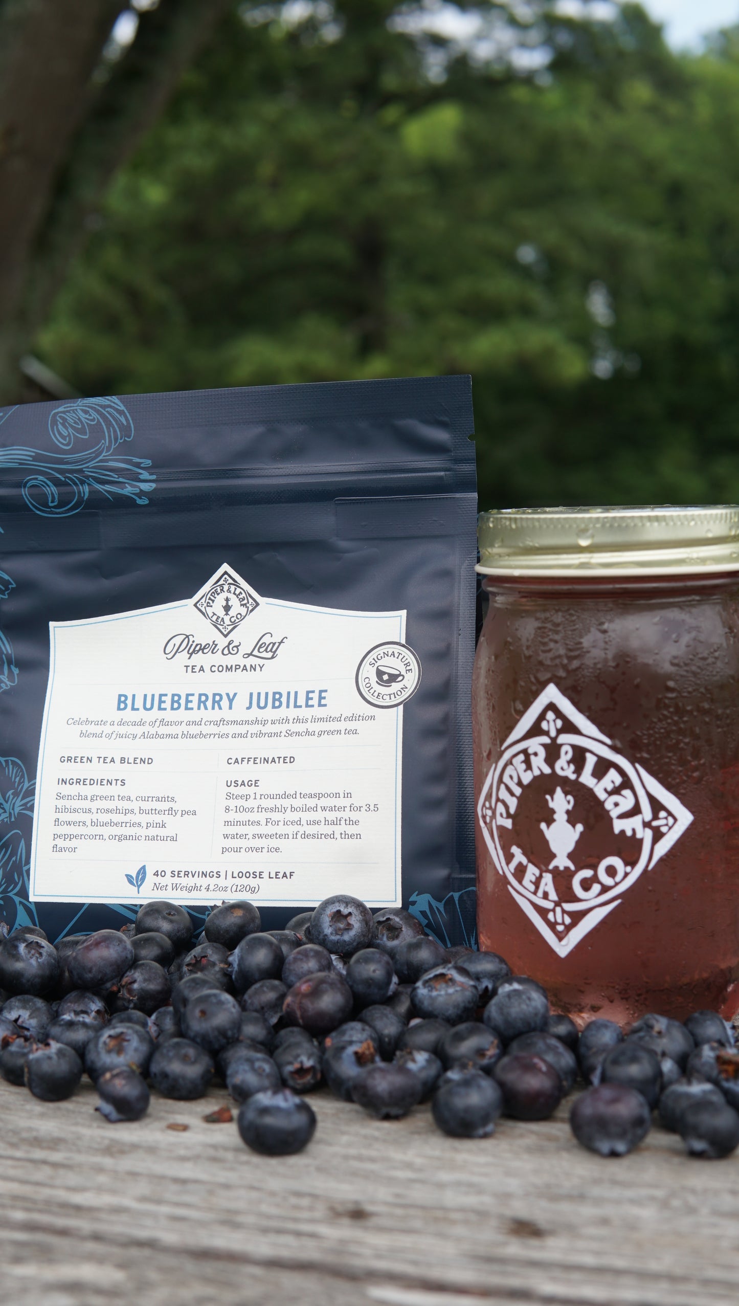 A bag of Piper and Leaf Tea Co. Blueberry Jubilee 40 Serving Loose Leaf, a jar of iced tea, and fresh blueberries are arranged on a wooden surface outdoors.