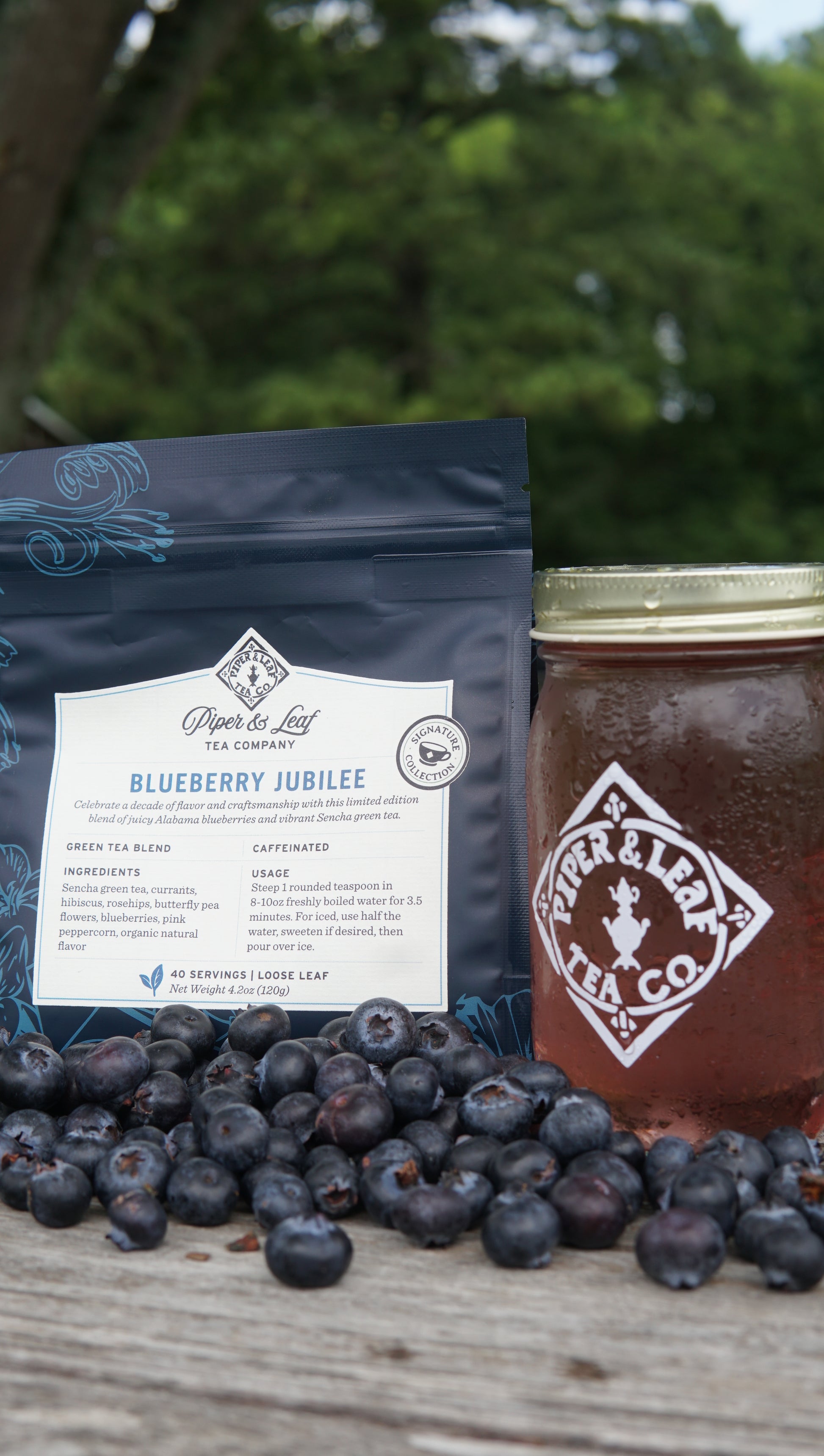 A bag of Piper and Leaf Tea Co. Blueberry Jubilee 40 Serving Loose Leaf, a jar of iced tea, and fresh blueberries are arranged on a wooden surface outdoors.