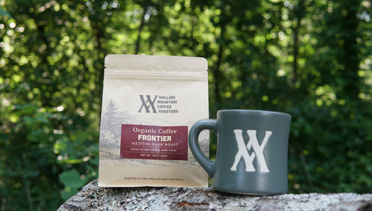 A 12oz Bag of Heritage Roast Coffee — Walling Mtn. and a gray mug sit outdoors, surrounded by green trees in the background.