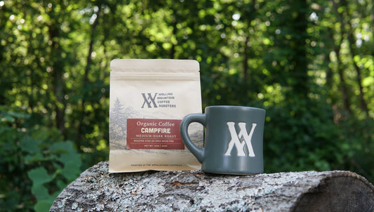 A 12oz Bag of Campfire Roast Coffee — Walling Mtn. and a green mug with a white logo rest on a log against a forest backdrop.