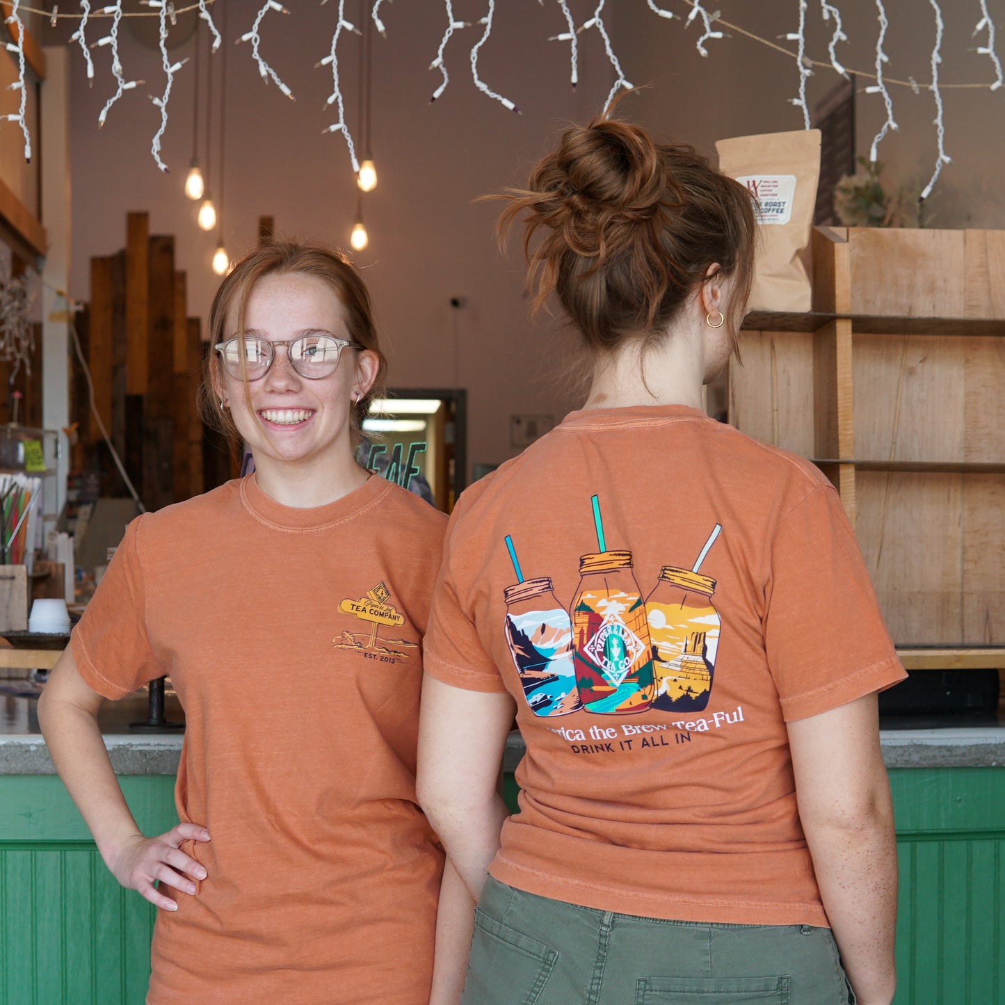 Two women wearing vibrant orange Piper & Leaf Tea Co. Westward Adventure Shirts stand in a cafe; one smiles at the camera while the other shows off the limited edition Adventure tee graphic on her back.