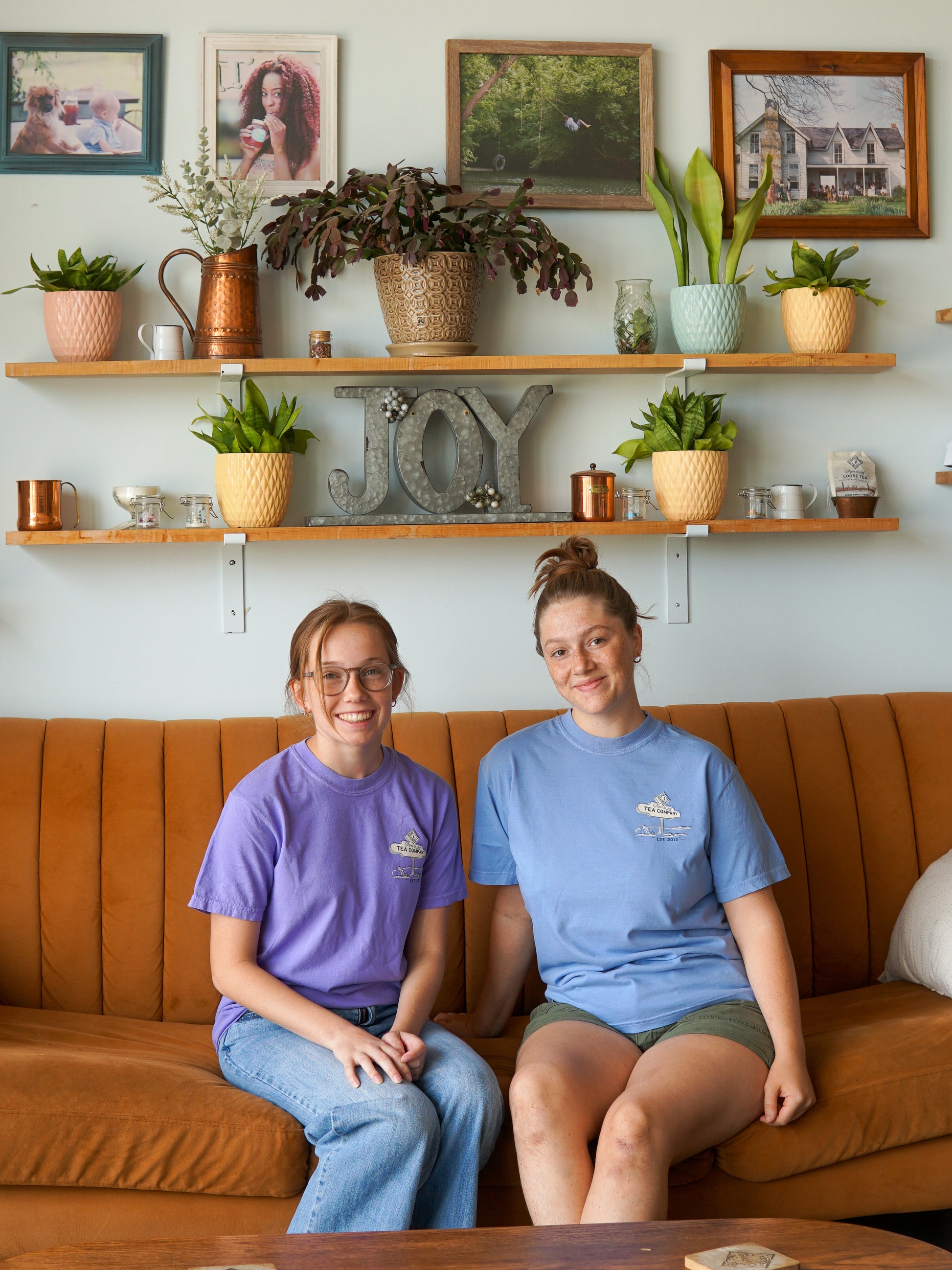 Two women wearing Nor'Easter Adventure T-shirts by Piper and Leaf Tea Co. sit on a brown couch in a living room decorated with plants, framed photos, and shelves holding decorative items, including a sign that reads "JOY.