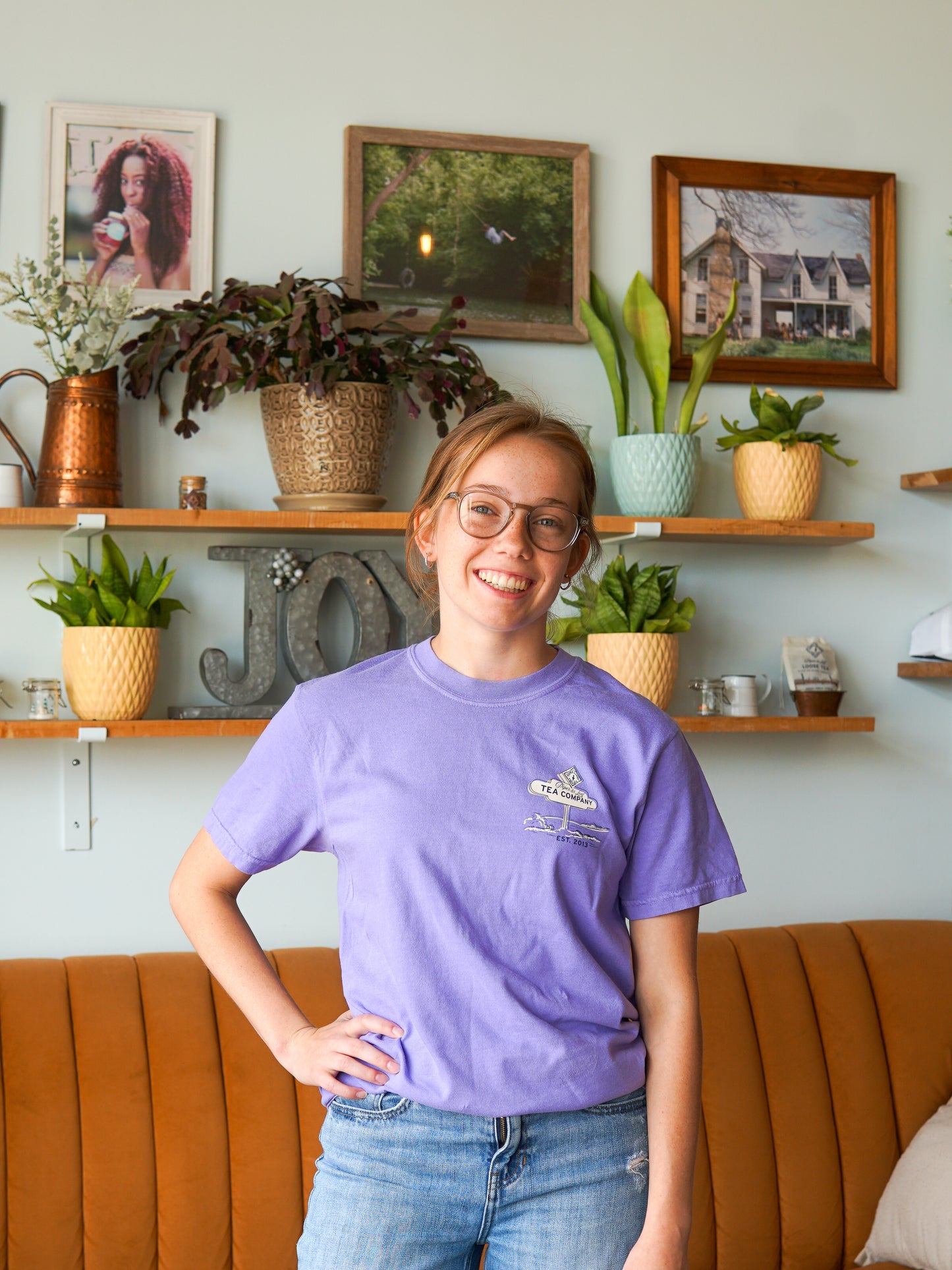 A young woman in glasses smiles in front of a tan couch, wearing the Nor'Easter Adventure T-shirt by Piper and Leaf Tea Co., with plants and framed pictures on shelves behind her.
