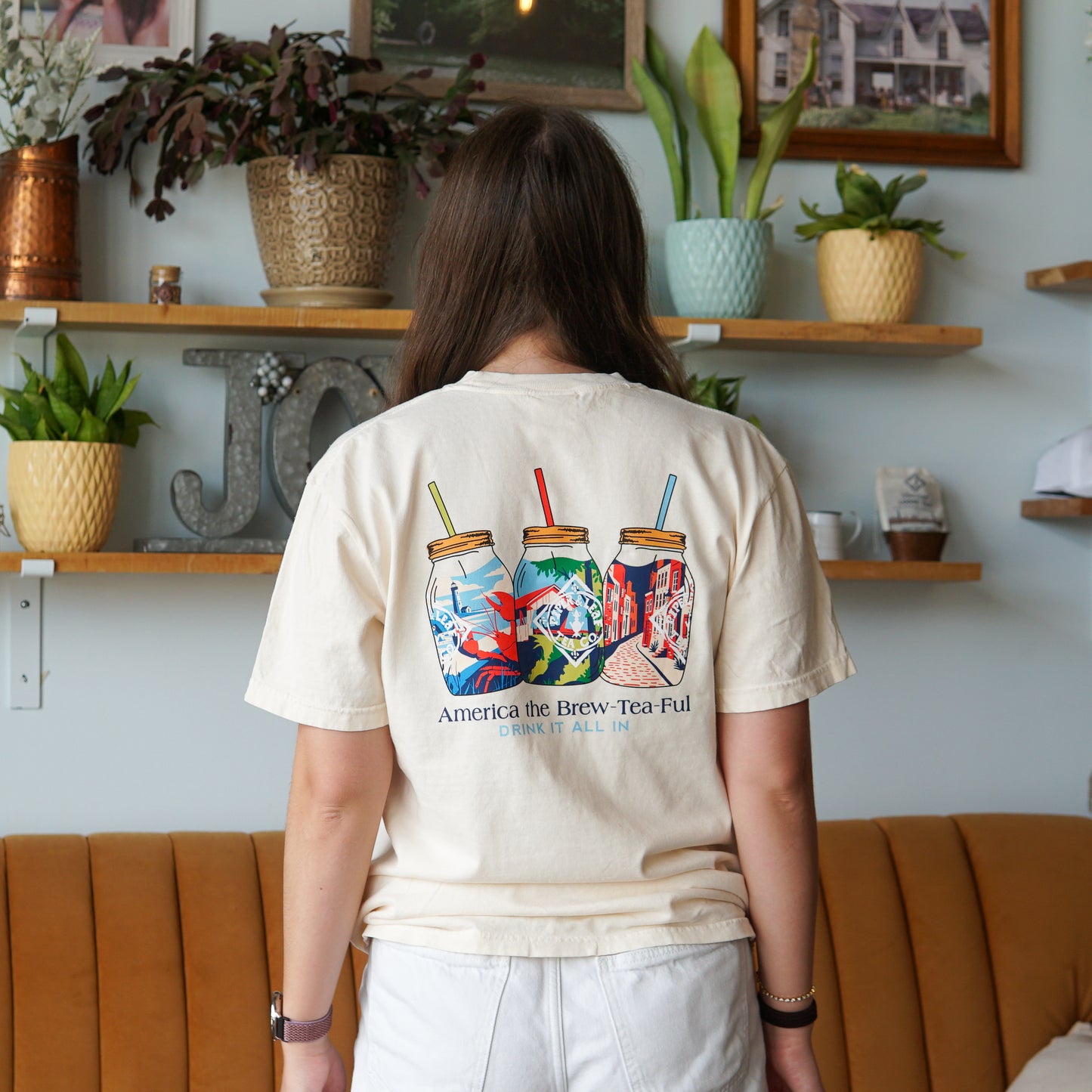 A person indoors near plants and shelves wears the Piper and Leaf Tea Co. Nor'Easter Adventure T-shirt, cream-colored with three mason jar illustrations and "America the Brew-Tea-Ful, Drink It All In" on the back.