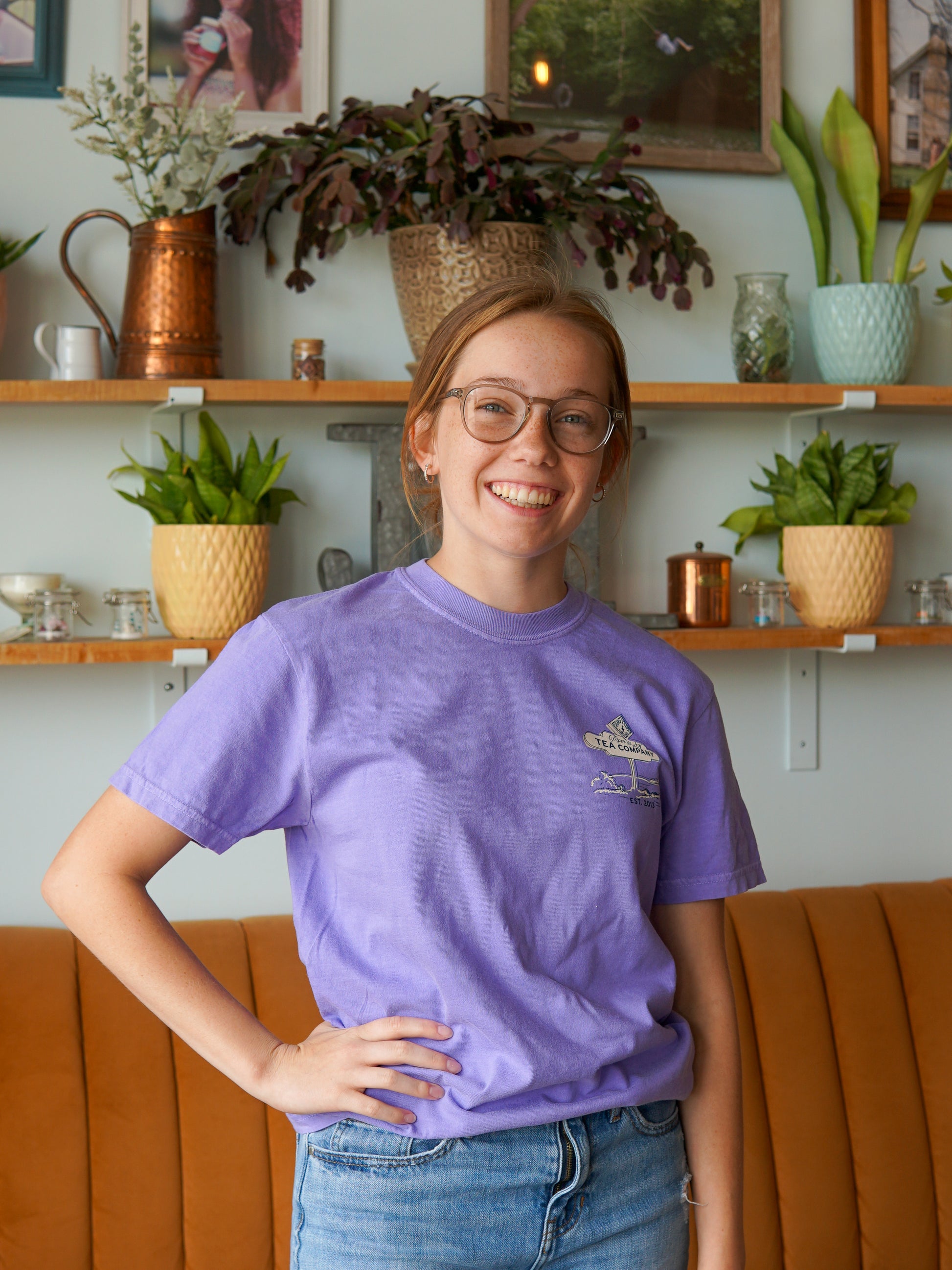 A young woman in glasses and a Nor'Easter Adventure T-shirt from Piper and Leaf Tea Co. smiles as she stands in front of shelves decorated with potted plants and ornaments.