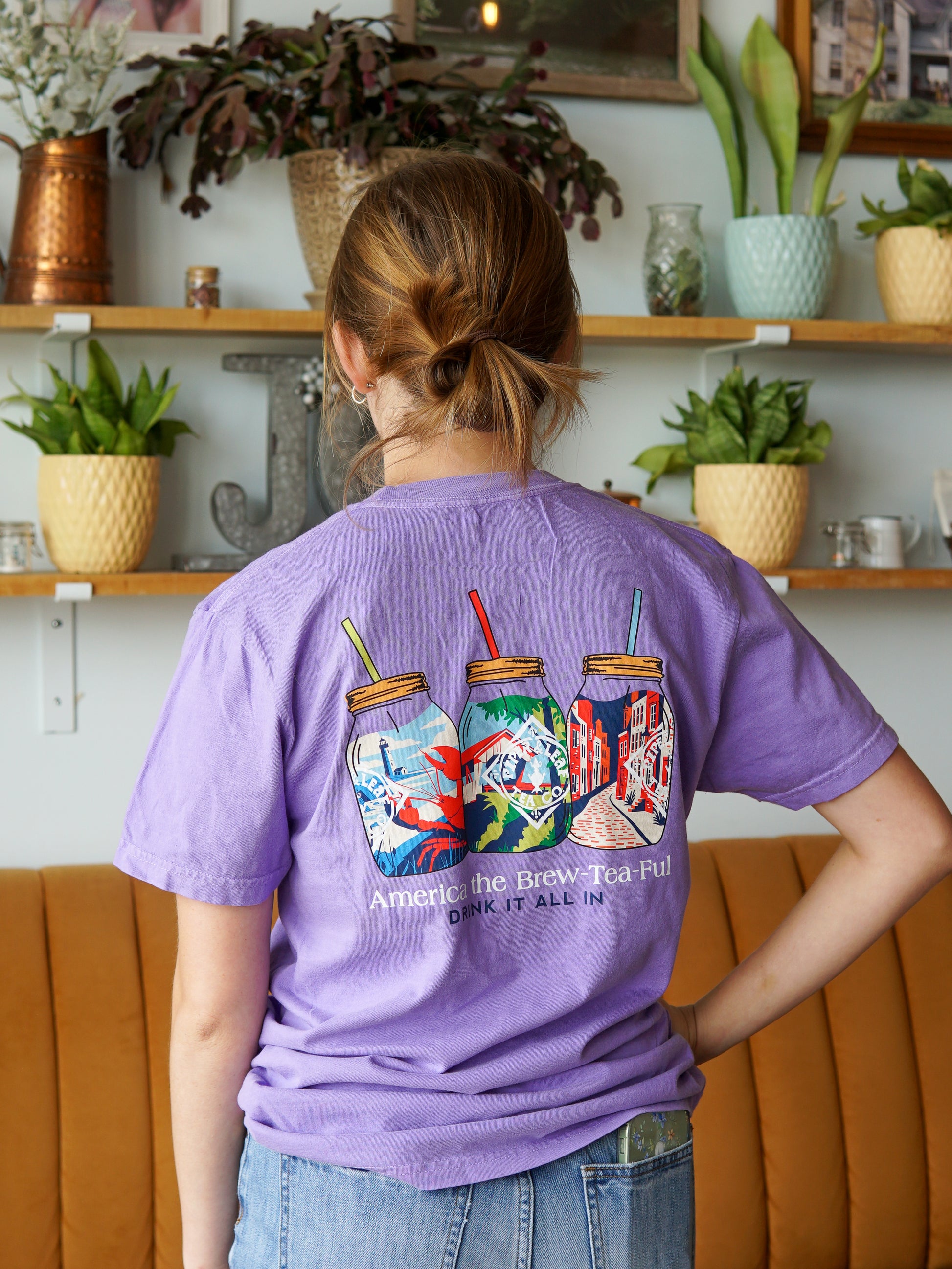 A person with light brown hair in a ponytail wears the Nor'Easter Adventure T-shirt by Piper and Leaf Tea Co., which is purple and features three illustrated mason jars and the text “America the Brew-Tea-Ful! DRINK IT ALL IN.” They stand by shelves of plants.