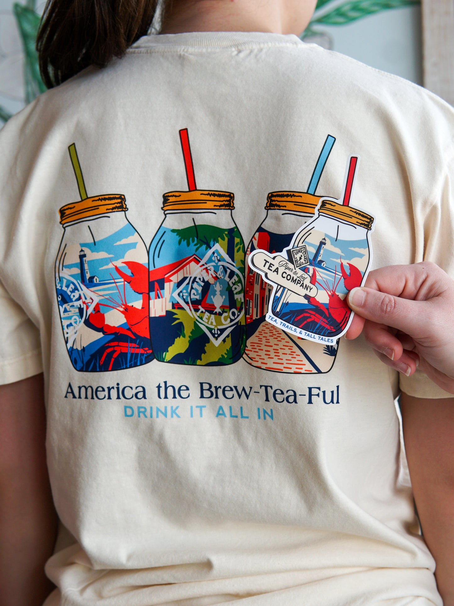 Someone wears the Piper and Leaf Tea Co. Nor'Easter Adventure T-shirt, featuring three mason jars with straws and the text "America the Brew-Tea-Ful, Drink It All In," while holding a matching sticker.