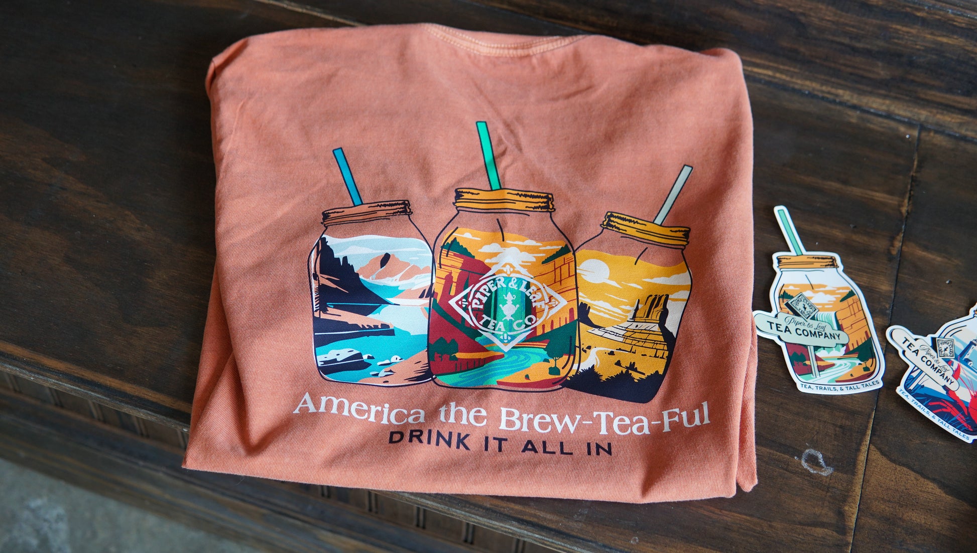 The Westward Adventure Shirt by Piper & Leaf Tea Co. is a comfort color orange tee featuring mason jar graphics, "America the Brew-Tea-Ful" text, and tea-themed stickers beside it—ideal for fans of limited edition Adventure tees.