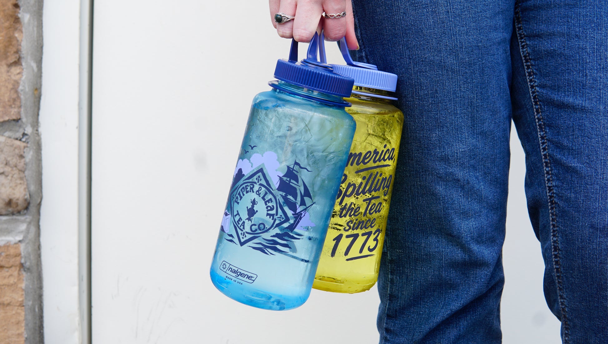 A person in jeans holds a blue bottle filled with ice and a Piper and Leaf Tea Co. "Spilling the Tea" Nalgene yellow water bottle with printed text.