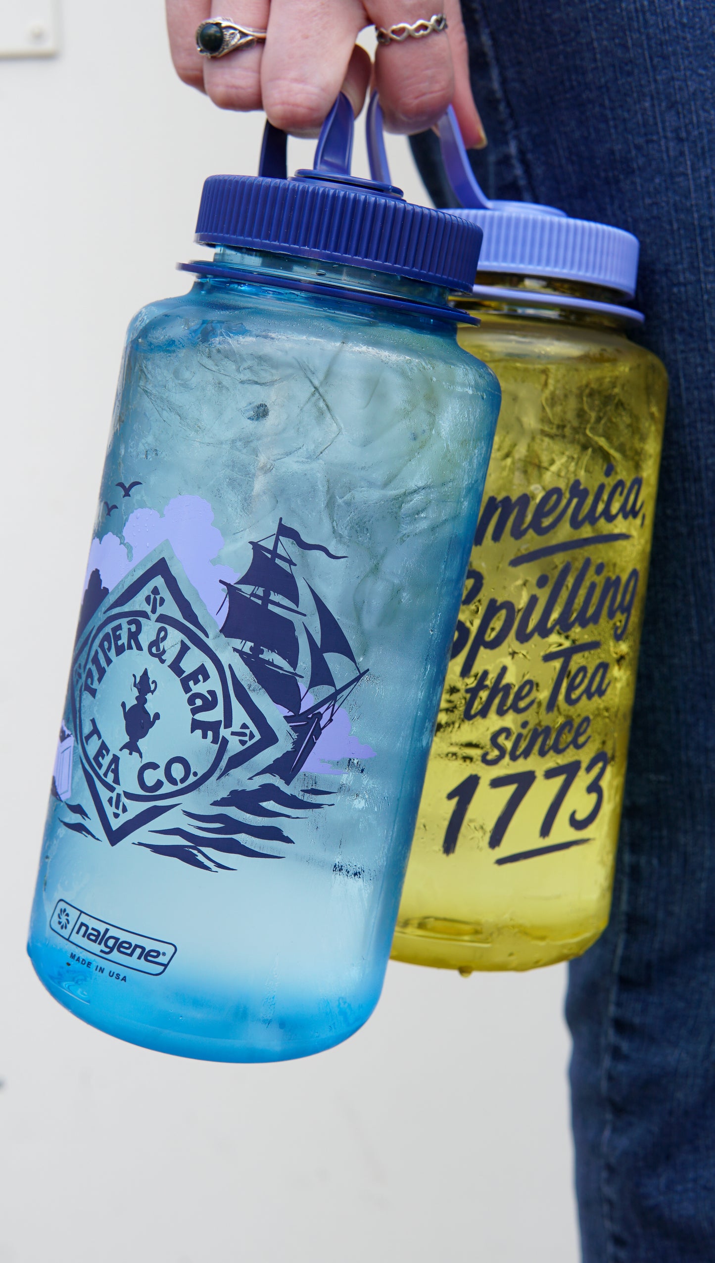 A person holds two large Nalgene bottles: a blue one with ice and a ship design, and Piper and Leaf Tea Co.’s yellow “Spilling the Tea Nalgene” featuring text about spilling tea since 1773.