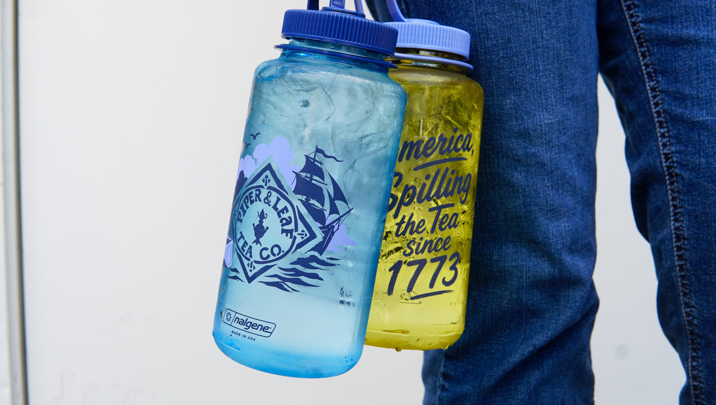 A person in jeans holds a blue bottle with a mountain logo and the yellow "Spilling the Tea Nalgene" by Piper and Leaf Tea Co., featuring text referencing tea and 1773.