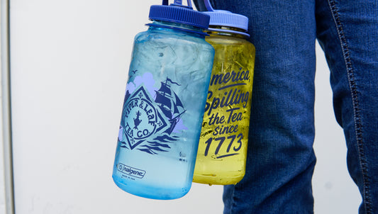 A person in jeans holds a blue bottle with a mountain logo and the yellow "Spilling the Tea Nalgene" by Piper and Leaf Tea Co., featuring text referencing tea and 1773.