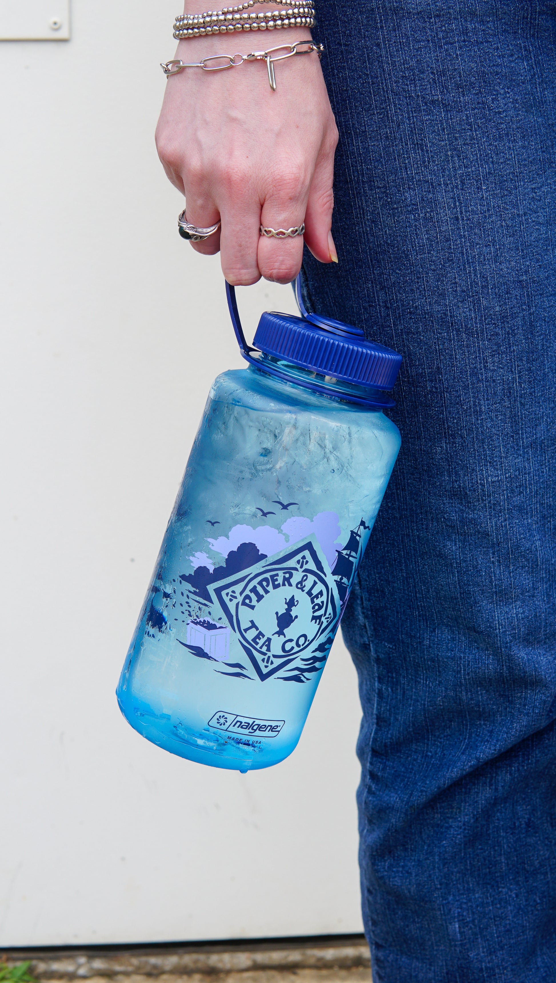 A person in blue jeans holds a large blue Spilling the Tea Nalgene water bottle by Piper and Leaf Tea Co., filled with ice water.
