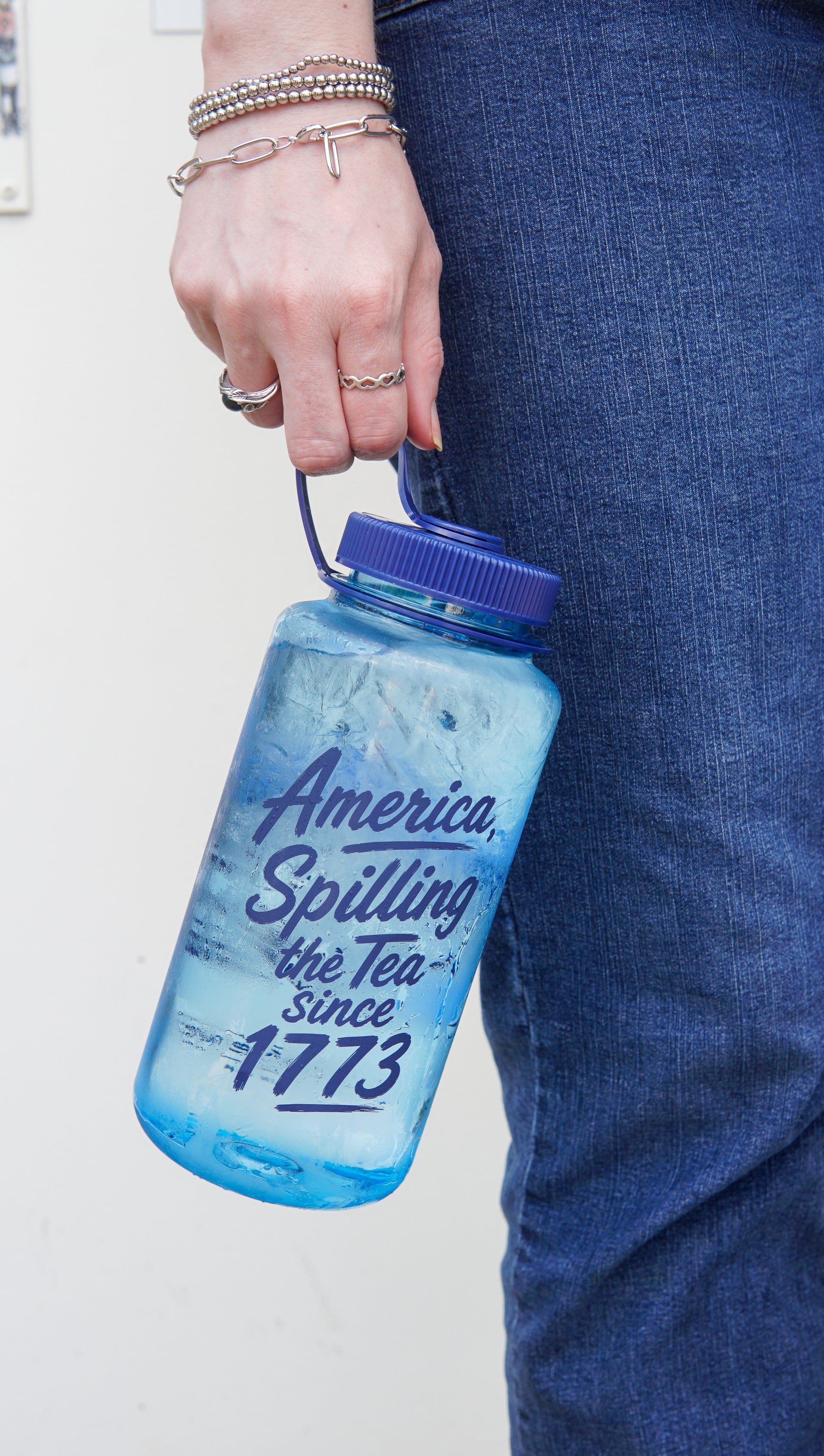 A person in blue jeans holds the Piper and Leaf Tea Co. "Spilling the Tea Nalgene" water bottle, featuring the text "America. Spilling the Tea since 1773.