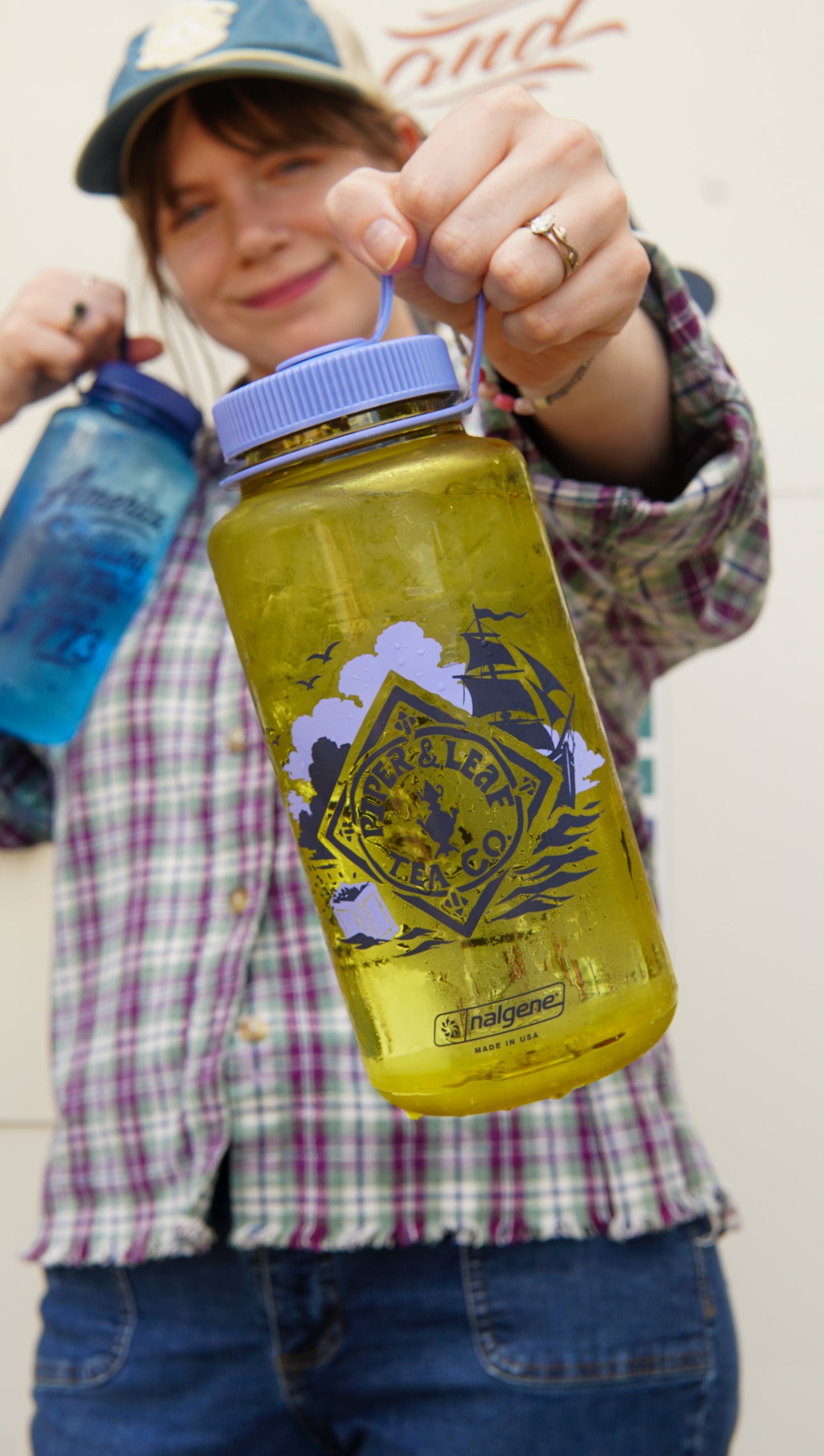 Person holding a clear yellow Spilling the Tea Nalgene by Piper and Leaf Tea Co., featuring a pirate-themed logo, with a blue bottle in the background, wearing a plaid shirt and hat.