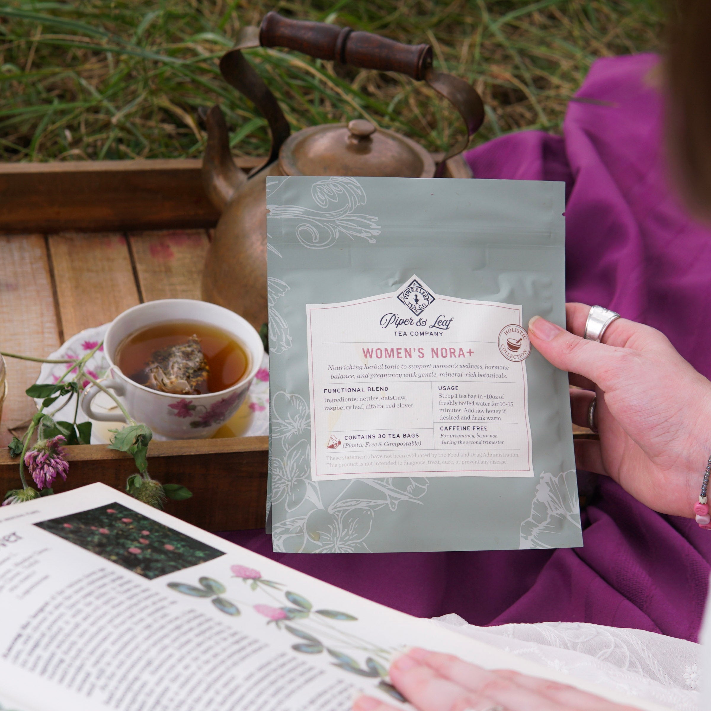 Someone holds a packet of Piper & Leaf Tea Co.’s Women’s NORA+—a tea blend for women’s wellness and hormone balance—beside a teacup, teapot, and open book on a wooden tray outdoors.
